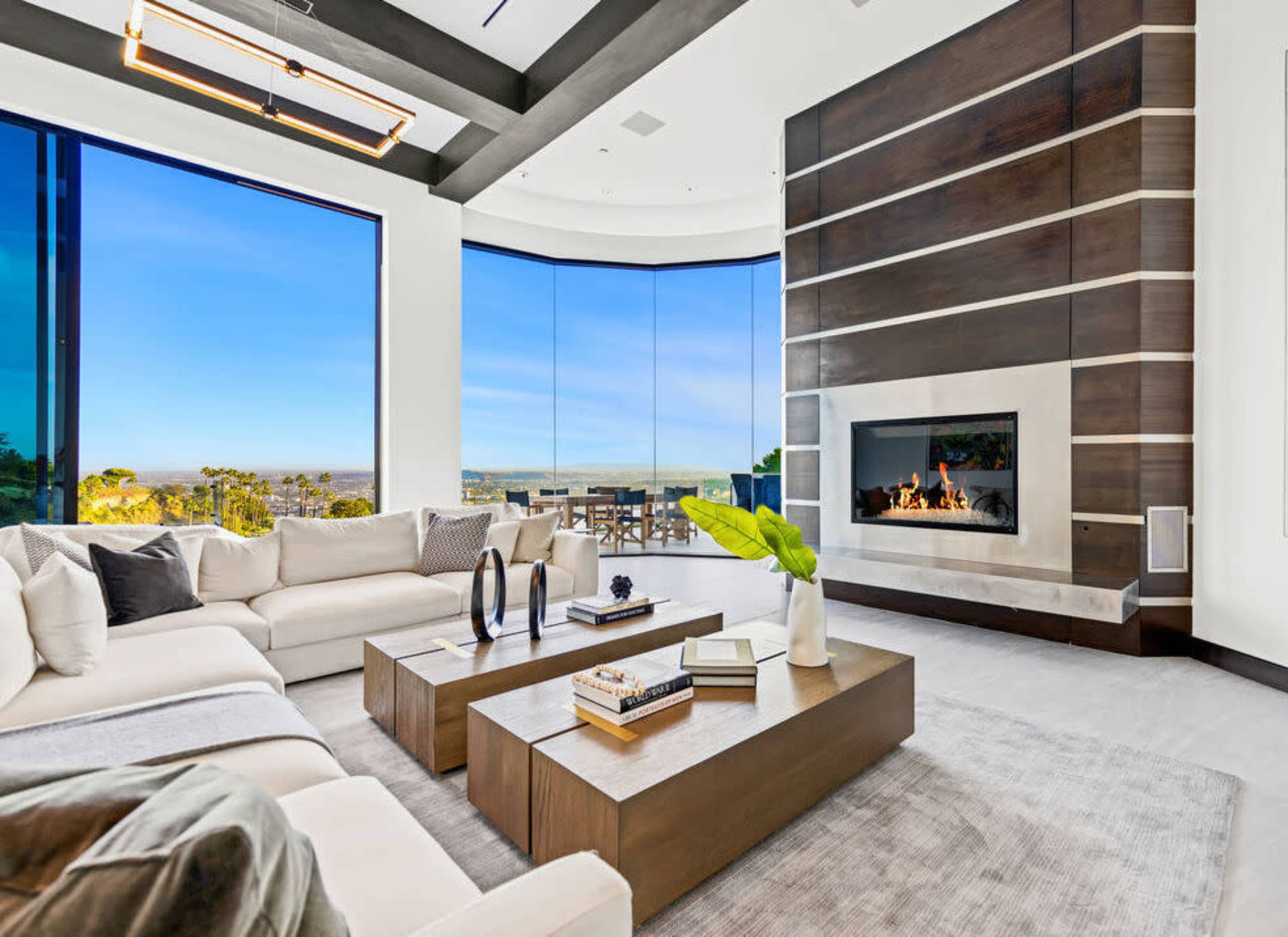The image shows a modern living room with large windows, a fireplace, and contemporary furniture arranged around a coffee table.