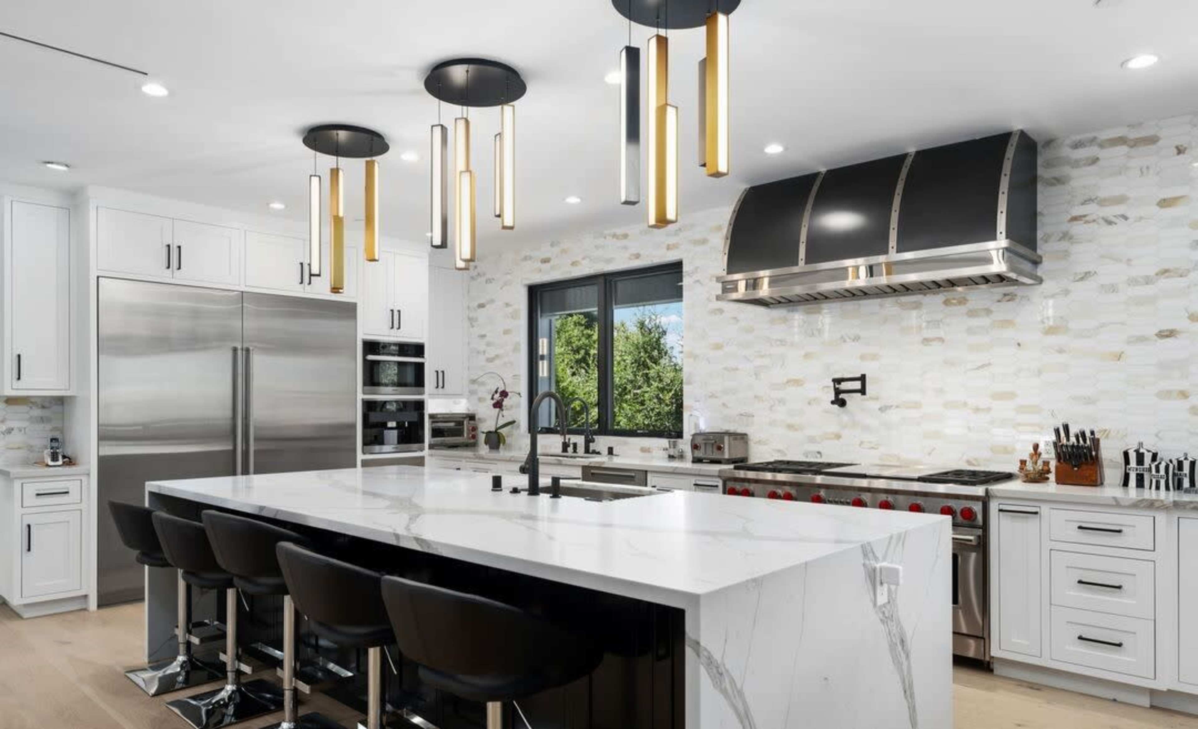 The modern kitchen features a large marble island with black bar stools, stainless steel appliances, and pendant lighting above.
