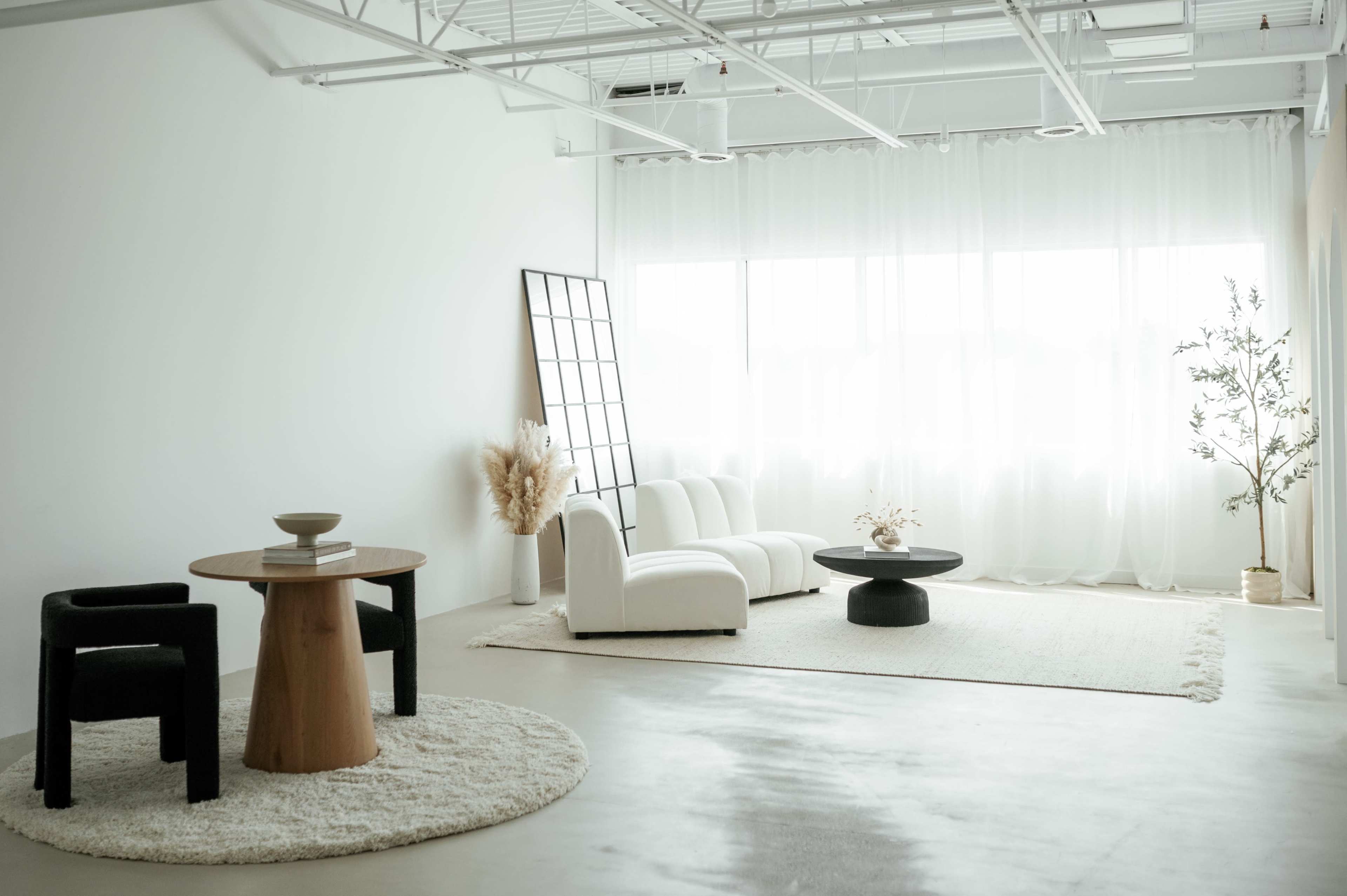 A minimally decorated living room with a white sofa, a round black coffee table, two chairs, and large windows covered by sheer curtains.