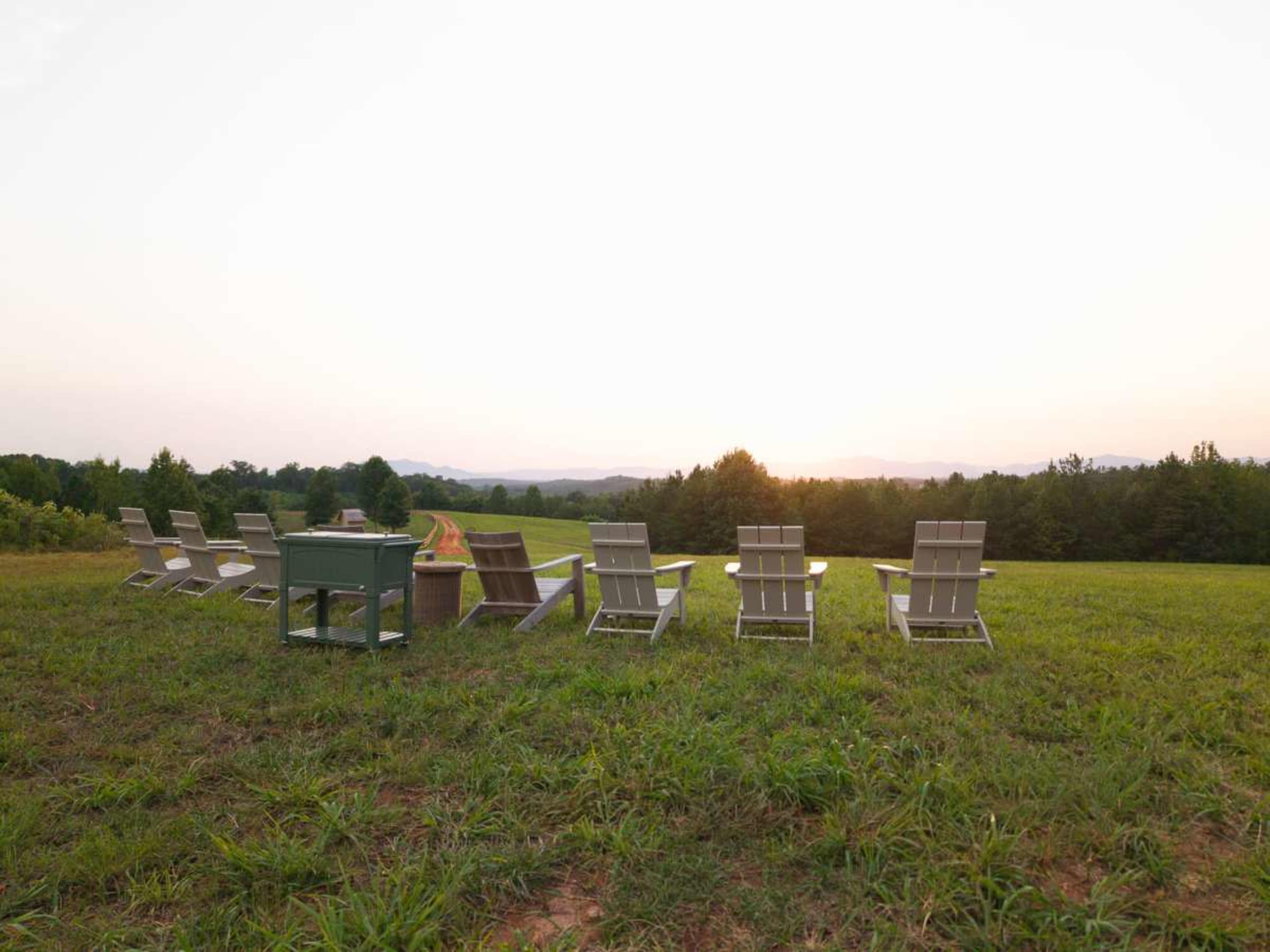 A row of white lounge chairs faces a distant sunset over a grassy field, with trees and mountains in the background.