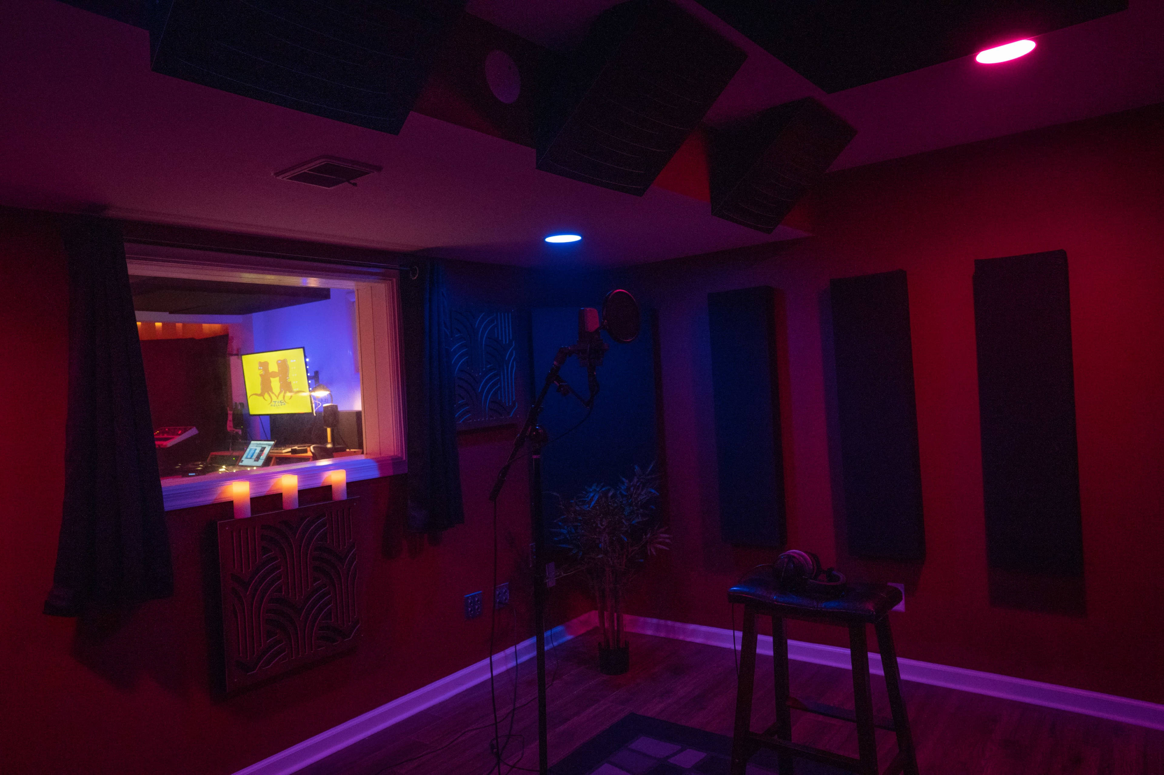 A dimly lit recording studio with a microphone on a stool, acoustic panels on the walls, and colorful lights illuminating the space.
