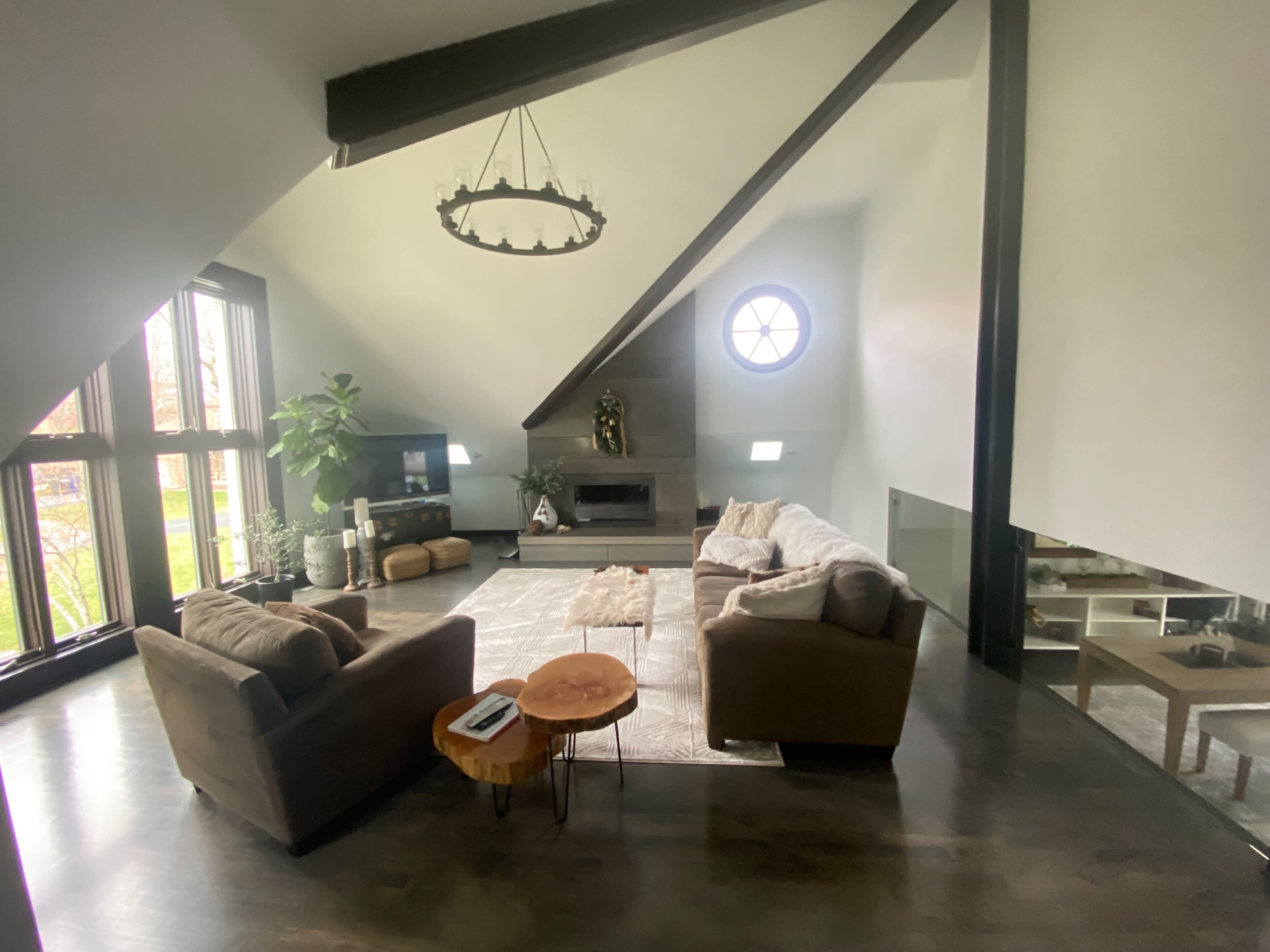 The image shows a spacious living room with a high ceiling, featuring a large sectional sofa, a coffee table, and a modern fireplace, alongside windows that allow natural light to enter.