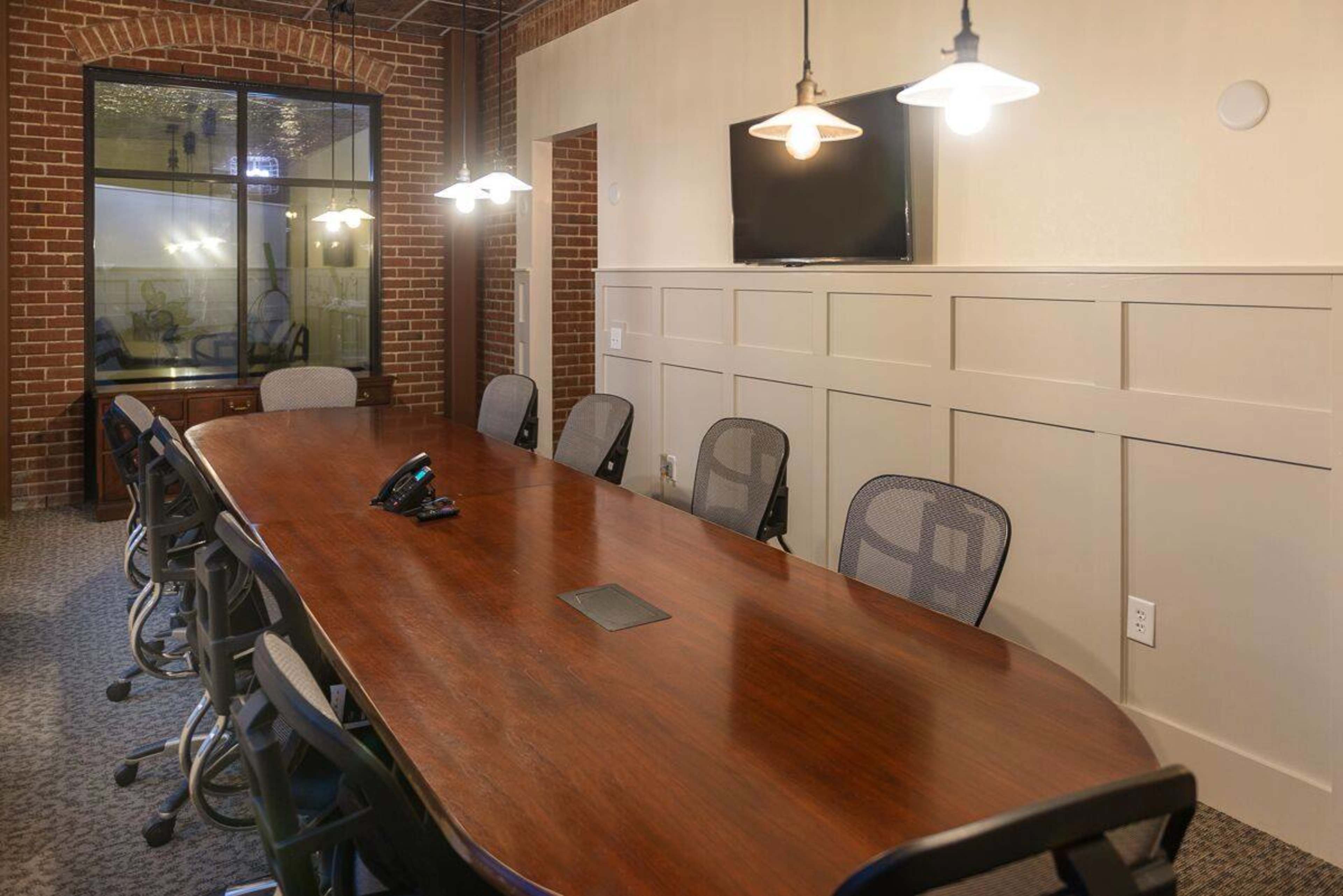 A long wooden conference table is surrounded by ergonomic chairs in a modern meeting room with a brick wall and a television mounted on one side.