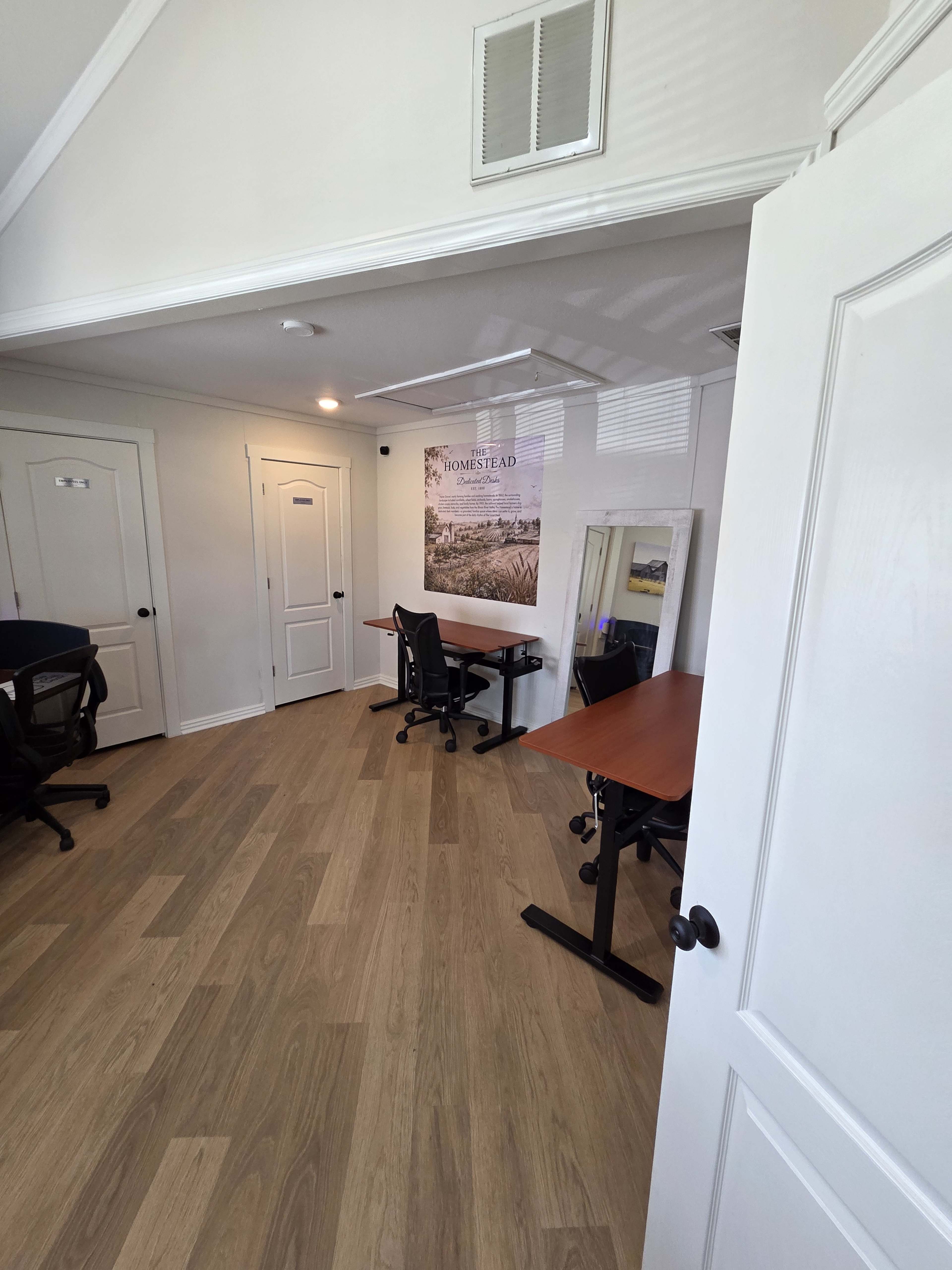 A small office space features two desks, a large mirror, and a poster on the wall, all illuminated by natural light.