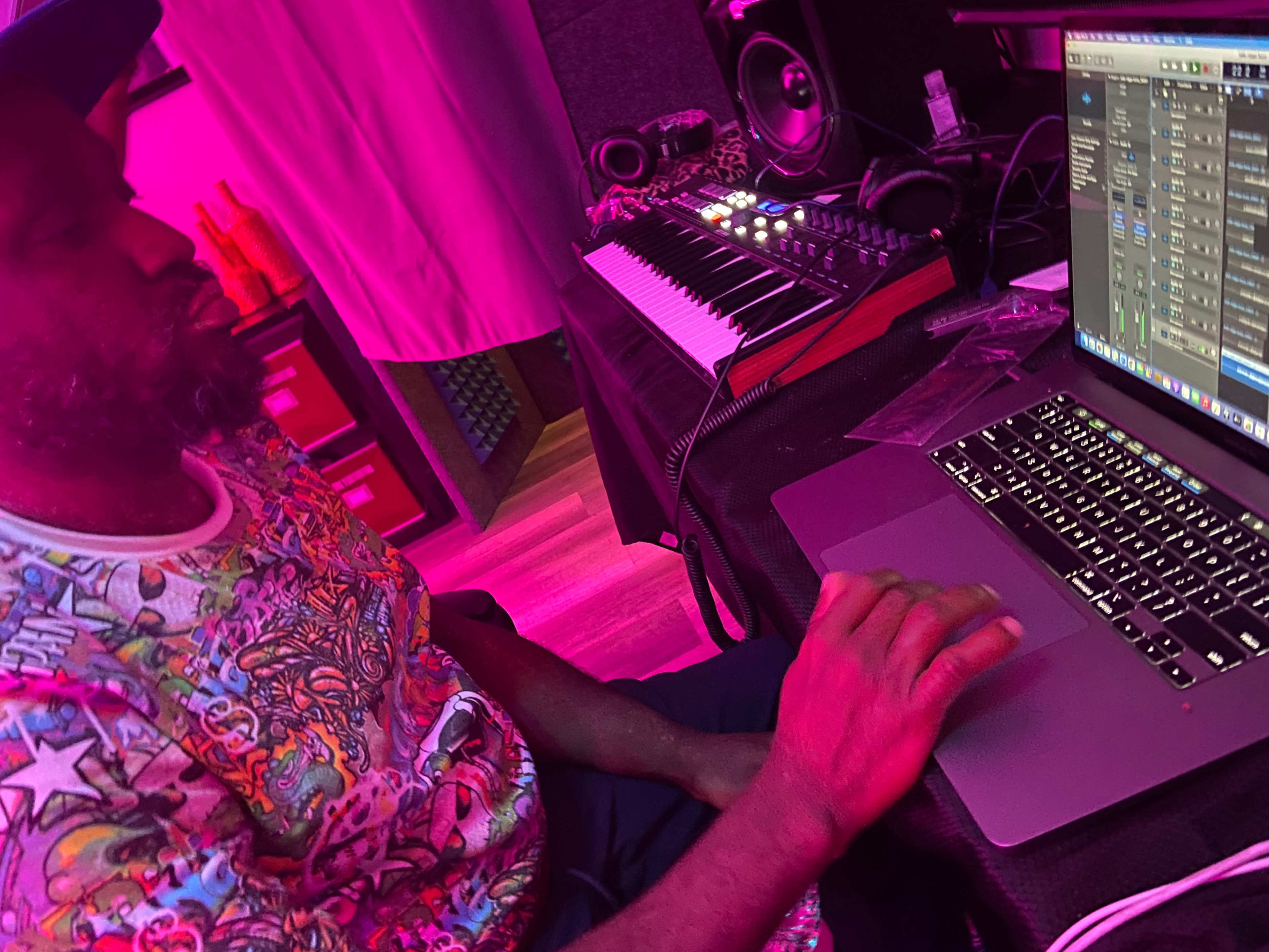 A person sits at a desk in a studio, working on a laptop and surrounded by music equipment and a keyboard in a dimly lit environment.