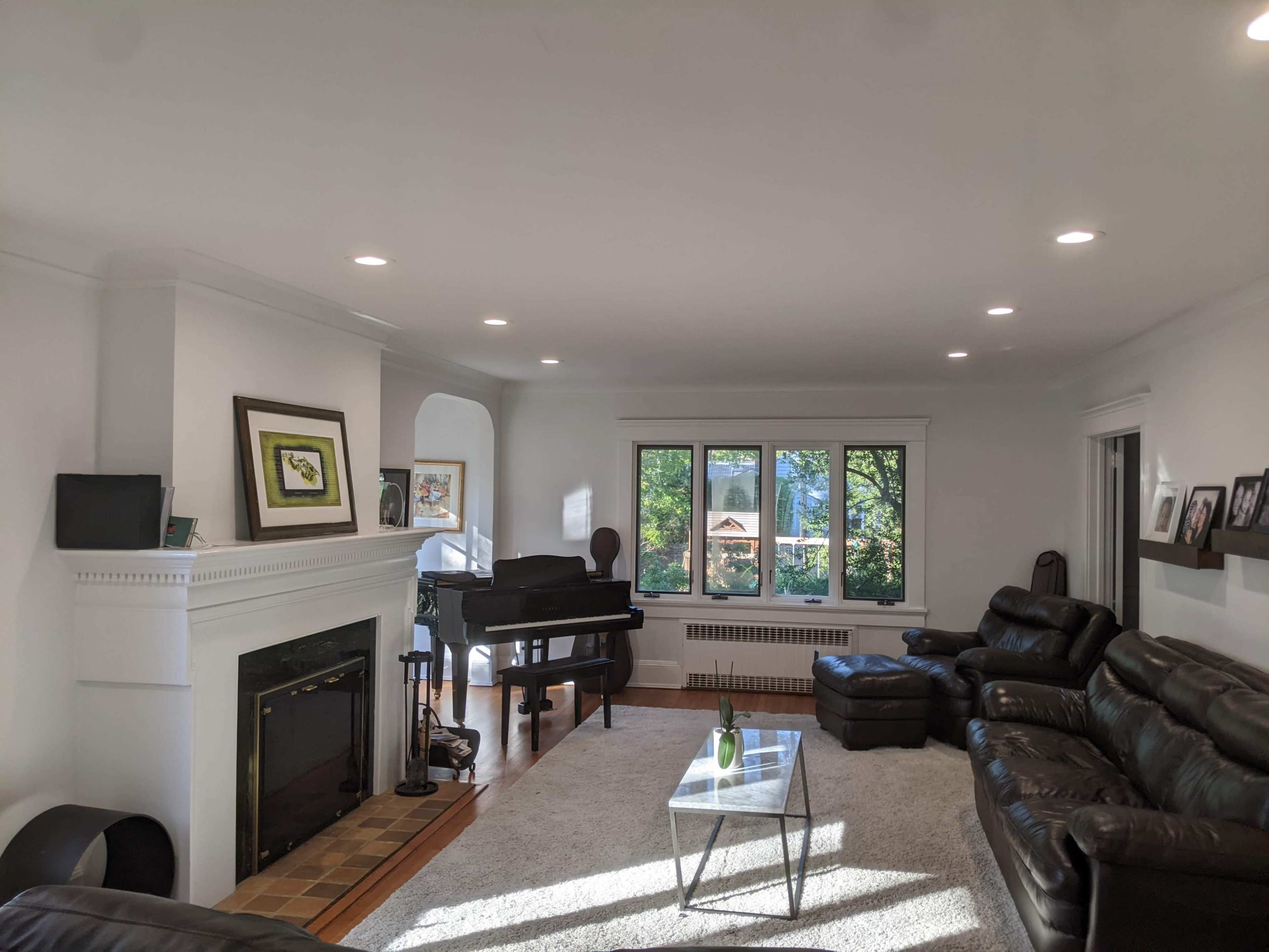 A spacious living room with dark leather sofas, a grand piano, a fireplace, and large windows overlooking greenery.
