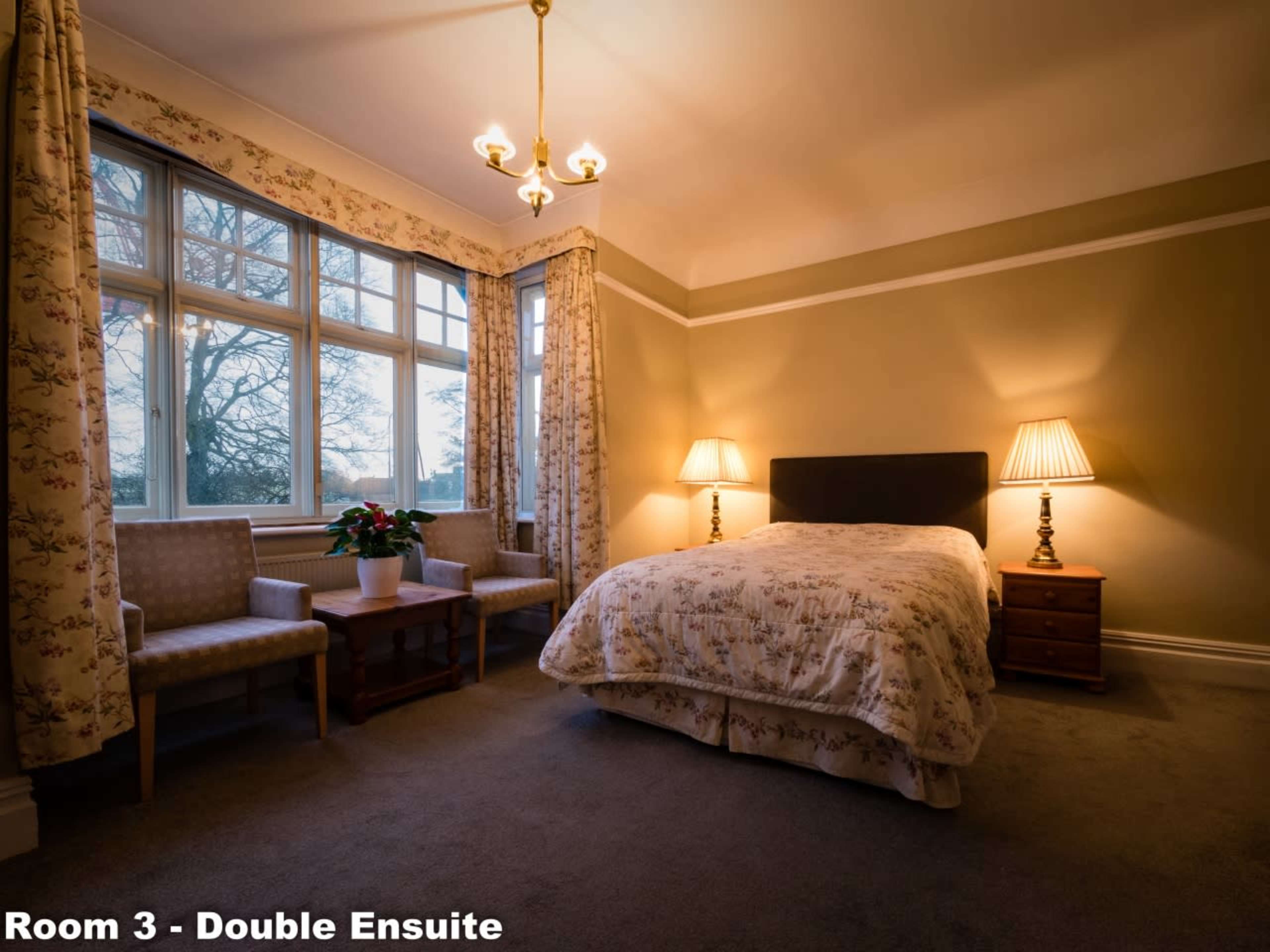 A well-lit bedroom featuring a double bed, two armchairs, a small table, and windows with curtains, set in a neutral-colored room.