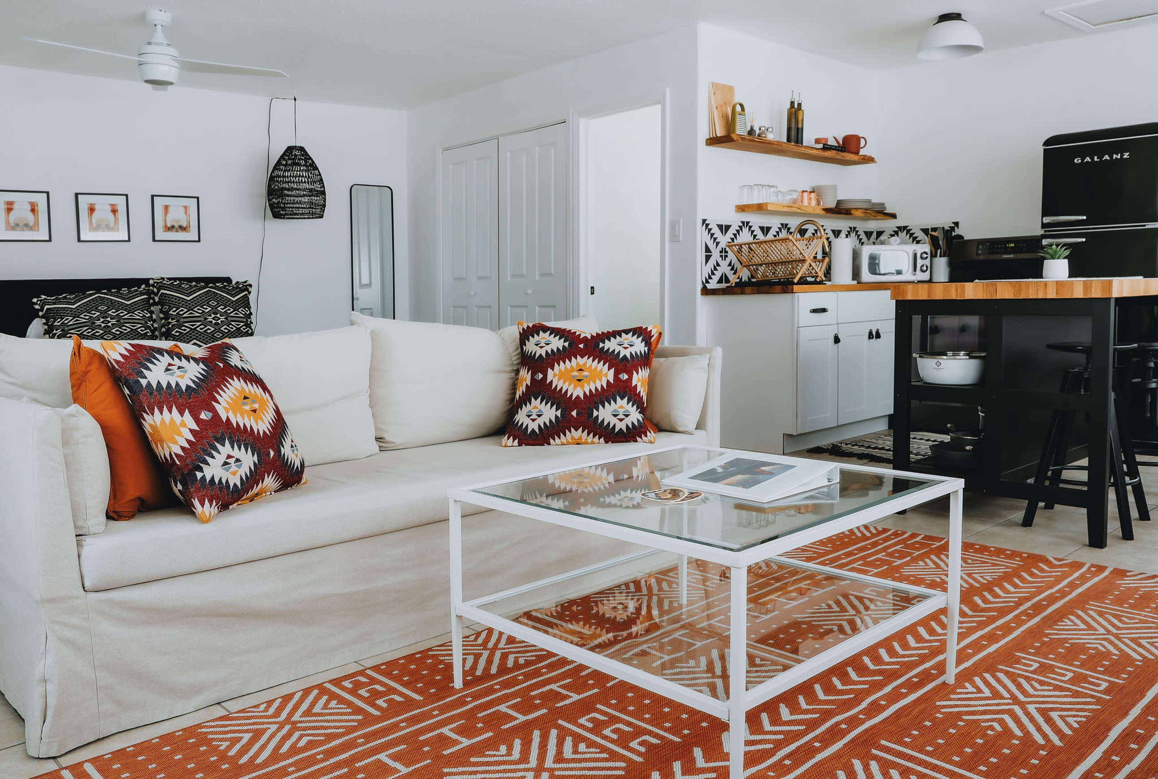 A modern living room with a white couch, a glass coffee table, a patterned rug, and a kitchen area in the background with black and white appliances.