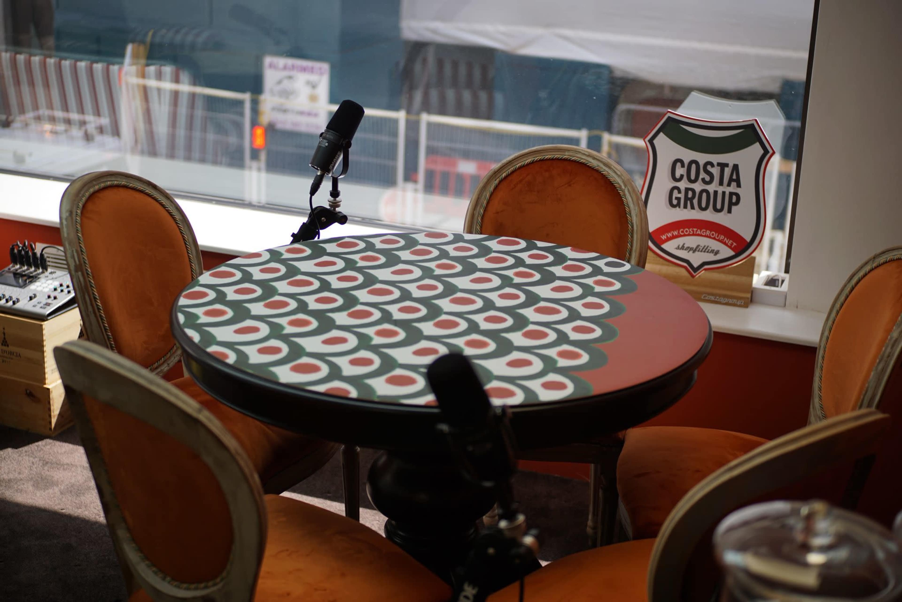 A round table with a patterned design is set up in front of a window, accompanied by orange chairs and two microphones on the table.