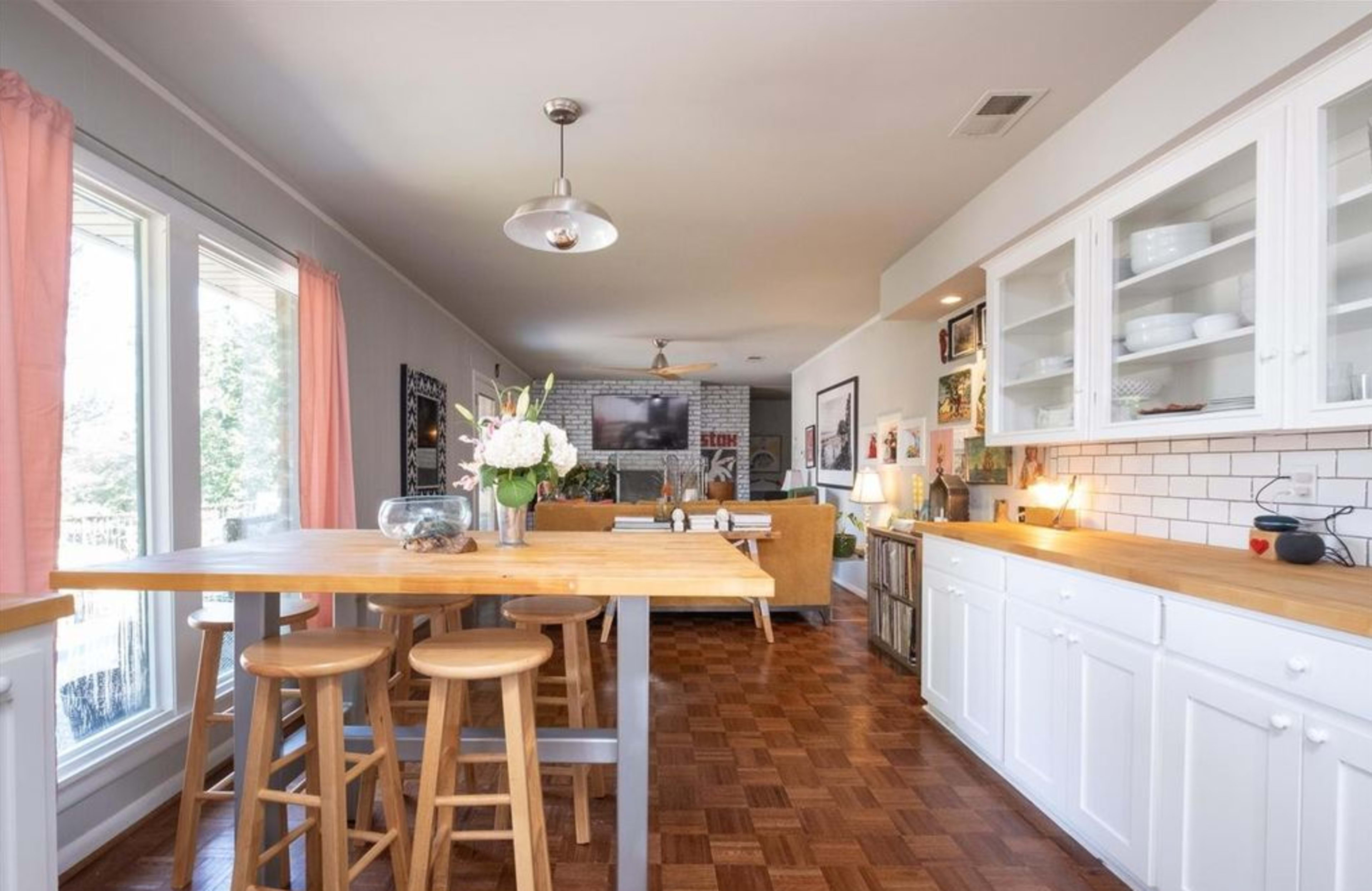 A modern open-concept kitchen and dining area featuring a wooden table with stools, white cabinetry, and large windows allowing natural light to fill the space.