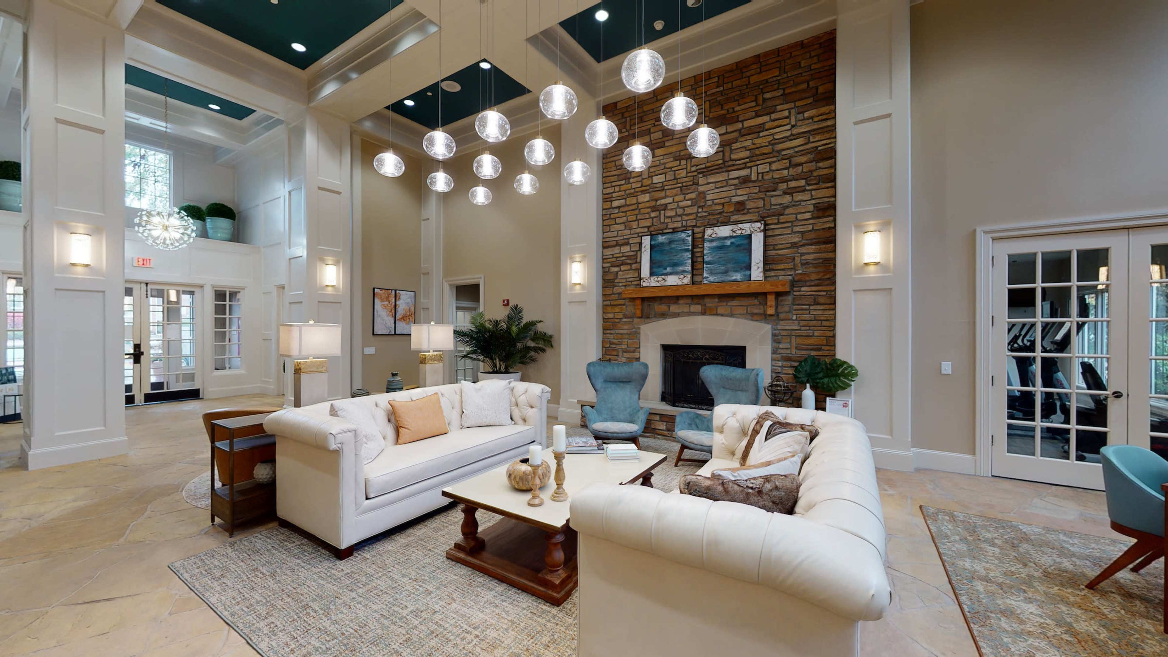 A spacious lobby area featuring a white sectional sofa, a wooden coffee table, a stone fireplace, and large pendant lights hanging from a high ceiling.