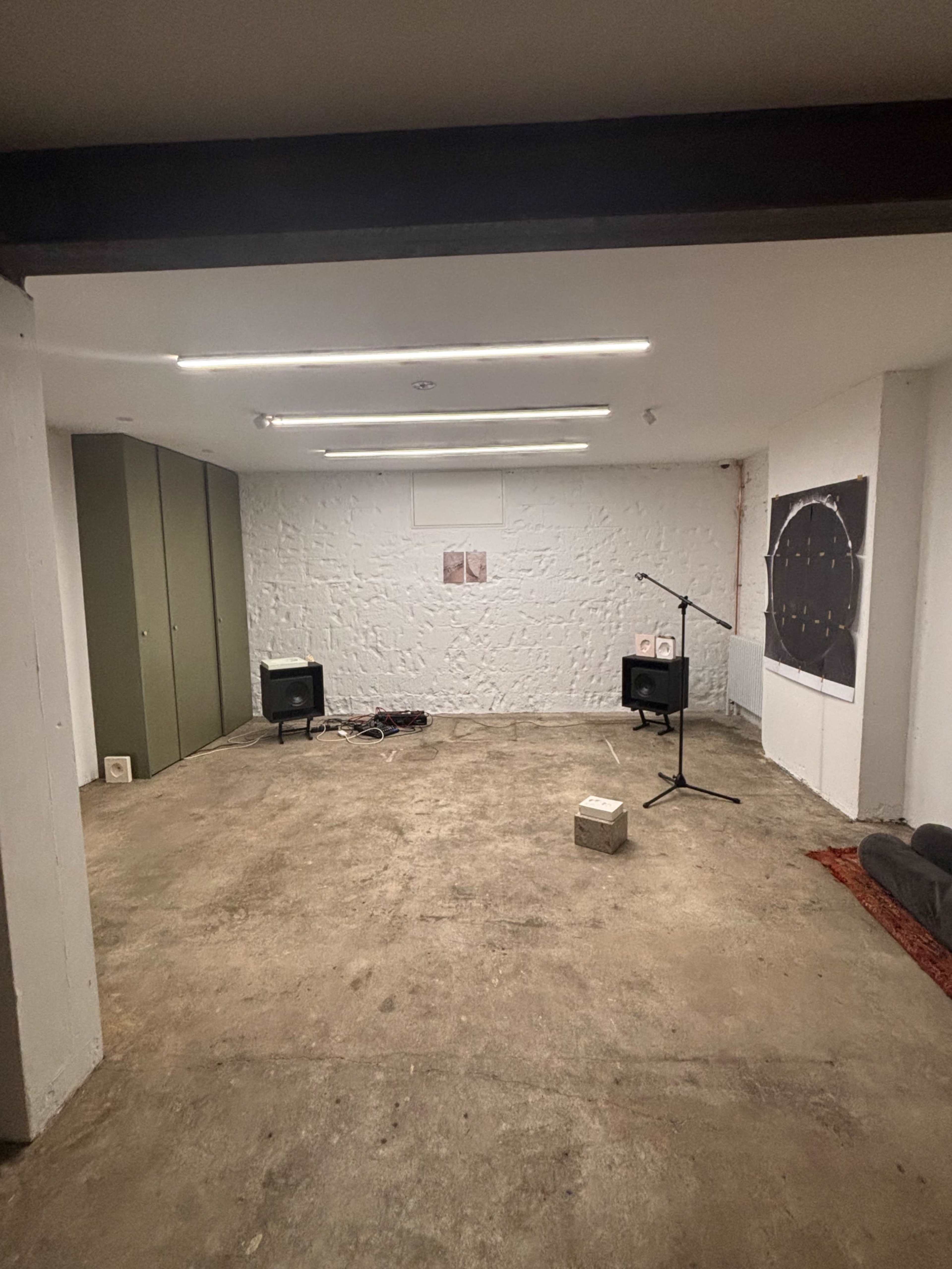 The image shows a sparse, empty room with a concrete floor, two speakers, a microphone on a stand, and minimal furnishings along the walls.