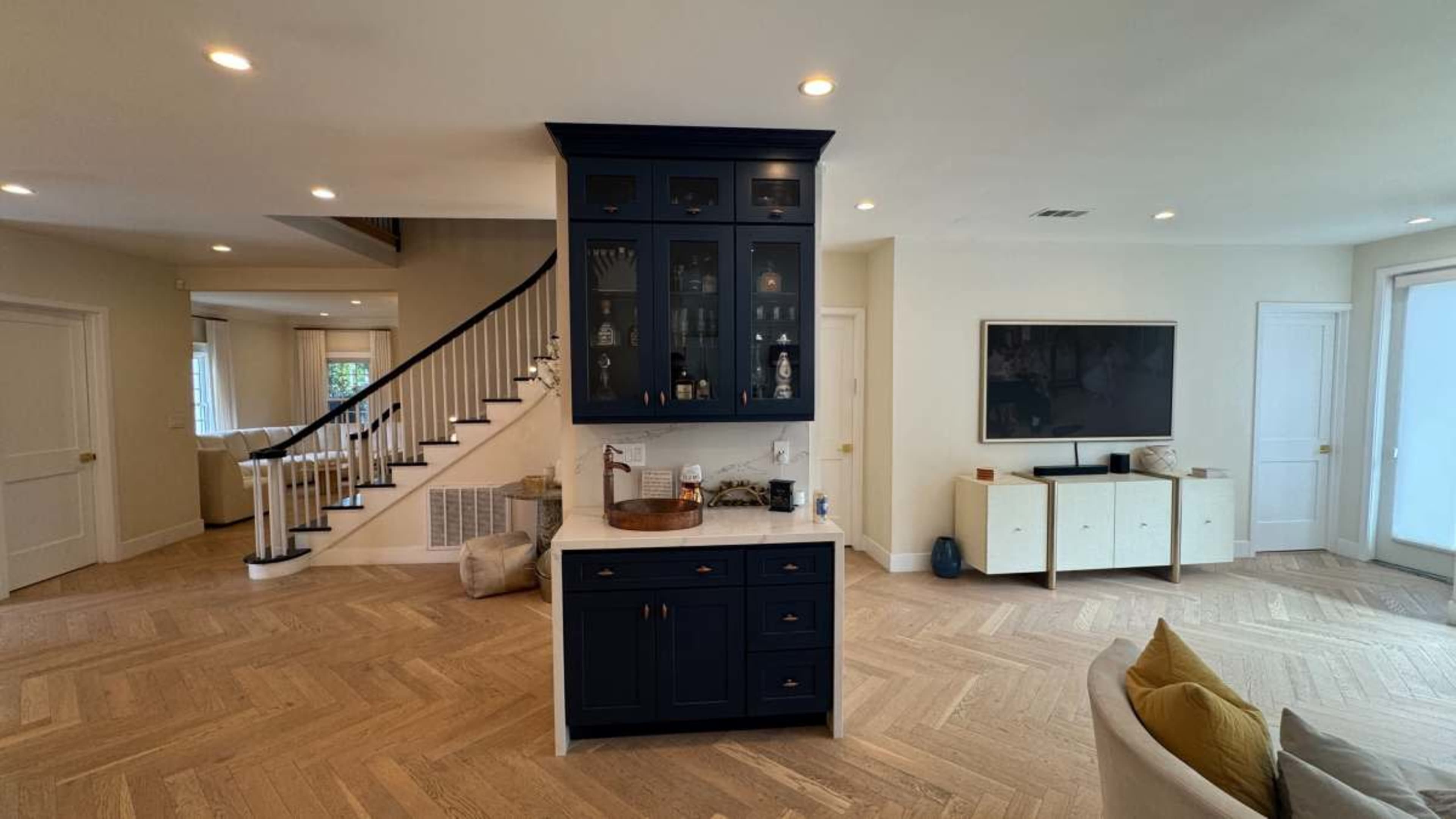 The image shows a modern kitchen with a dark blue island and cabinetry, surrounded by light-colored walls and hardwood flooring, leading to a staircase in the background.