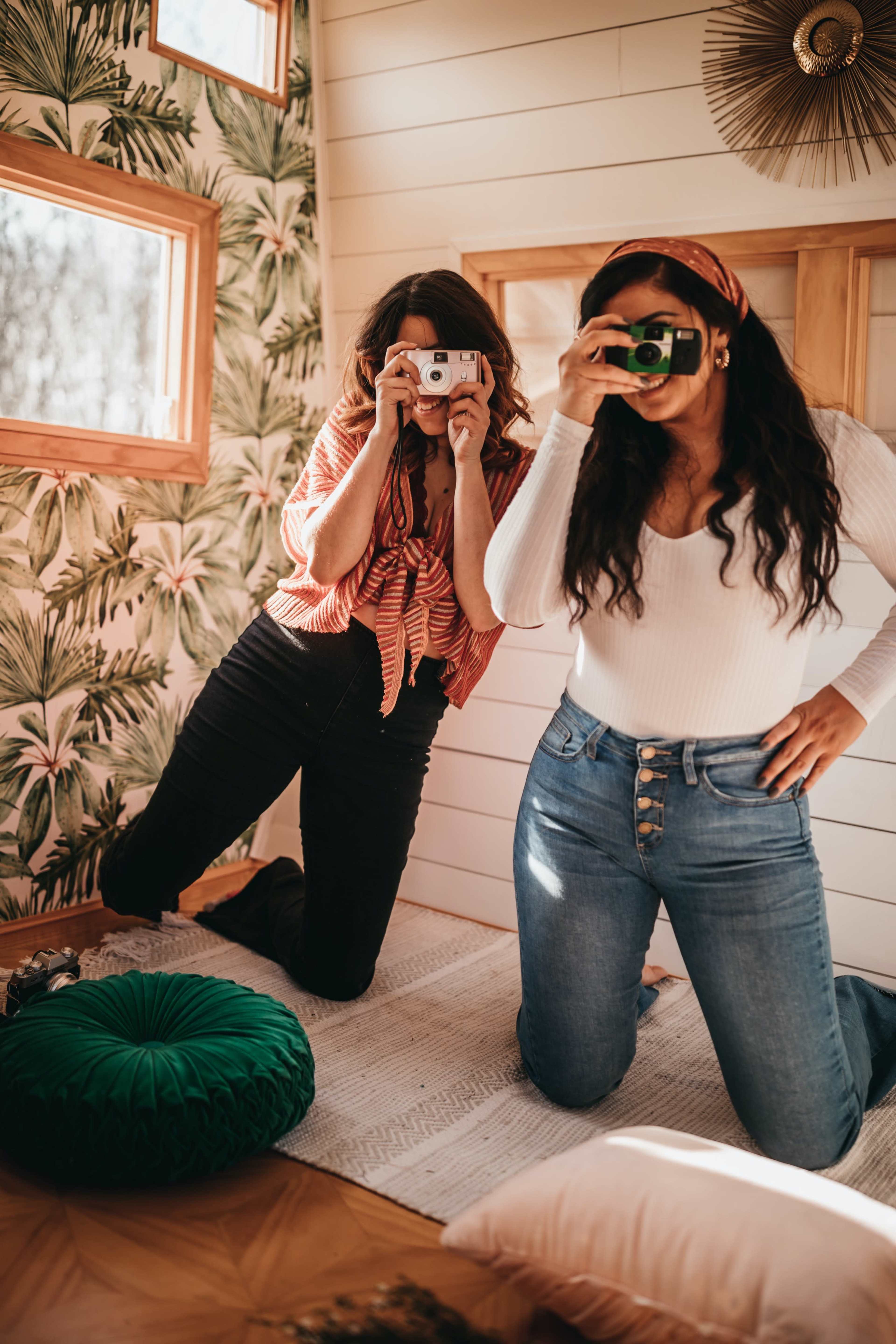 Two women kneel on a rug in a decorated interior, each holding a camera and smiling at the viewer.