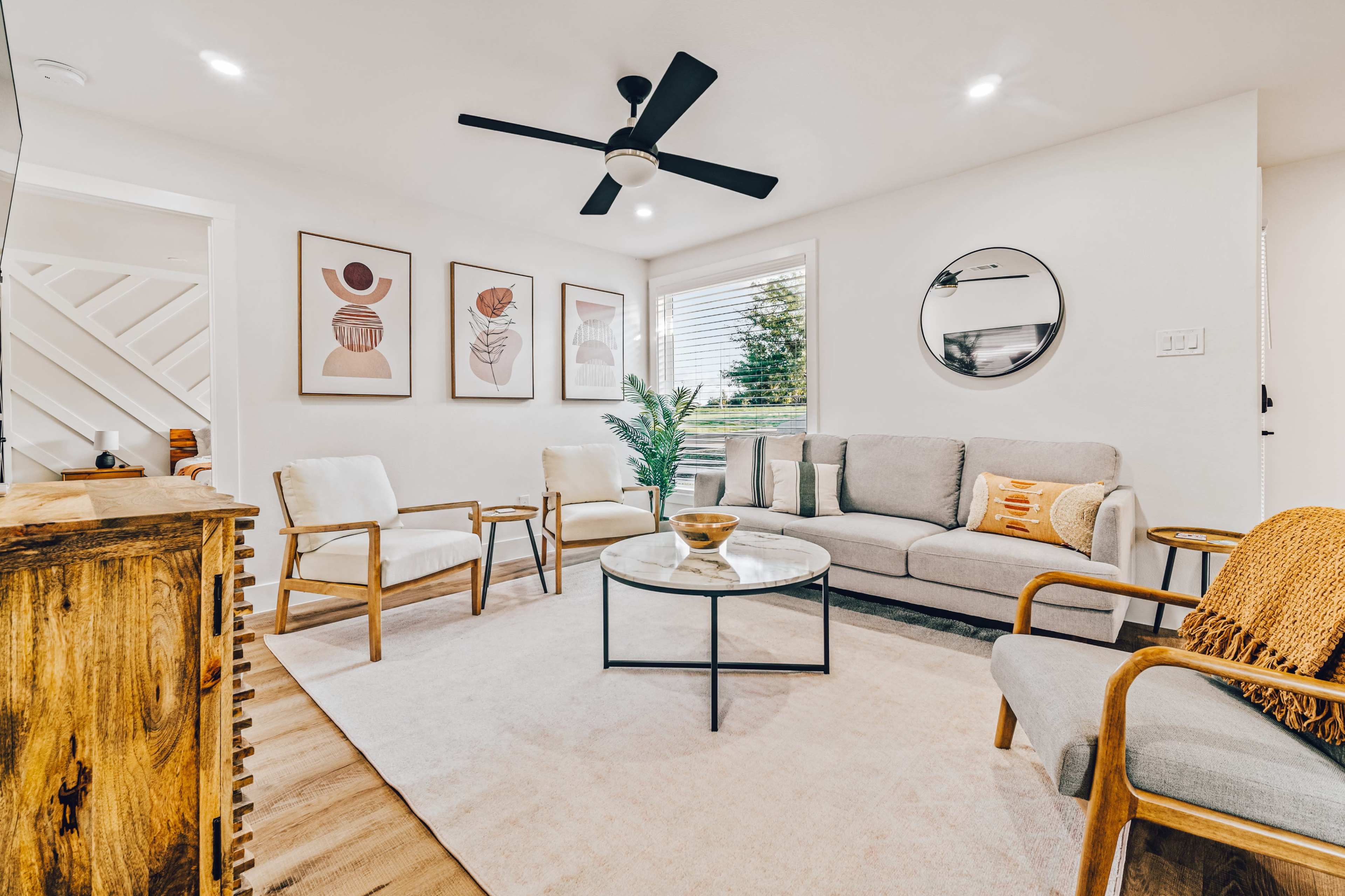 A modern living room with a sofa, two armchairs, a round glass coffee table, framed artwork on the walls, and a ceiling fan.