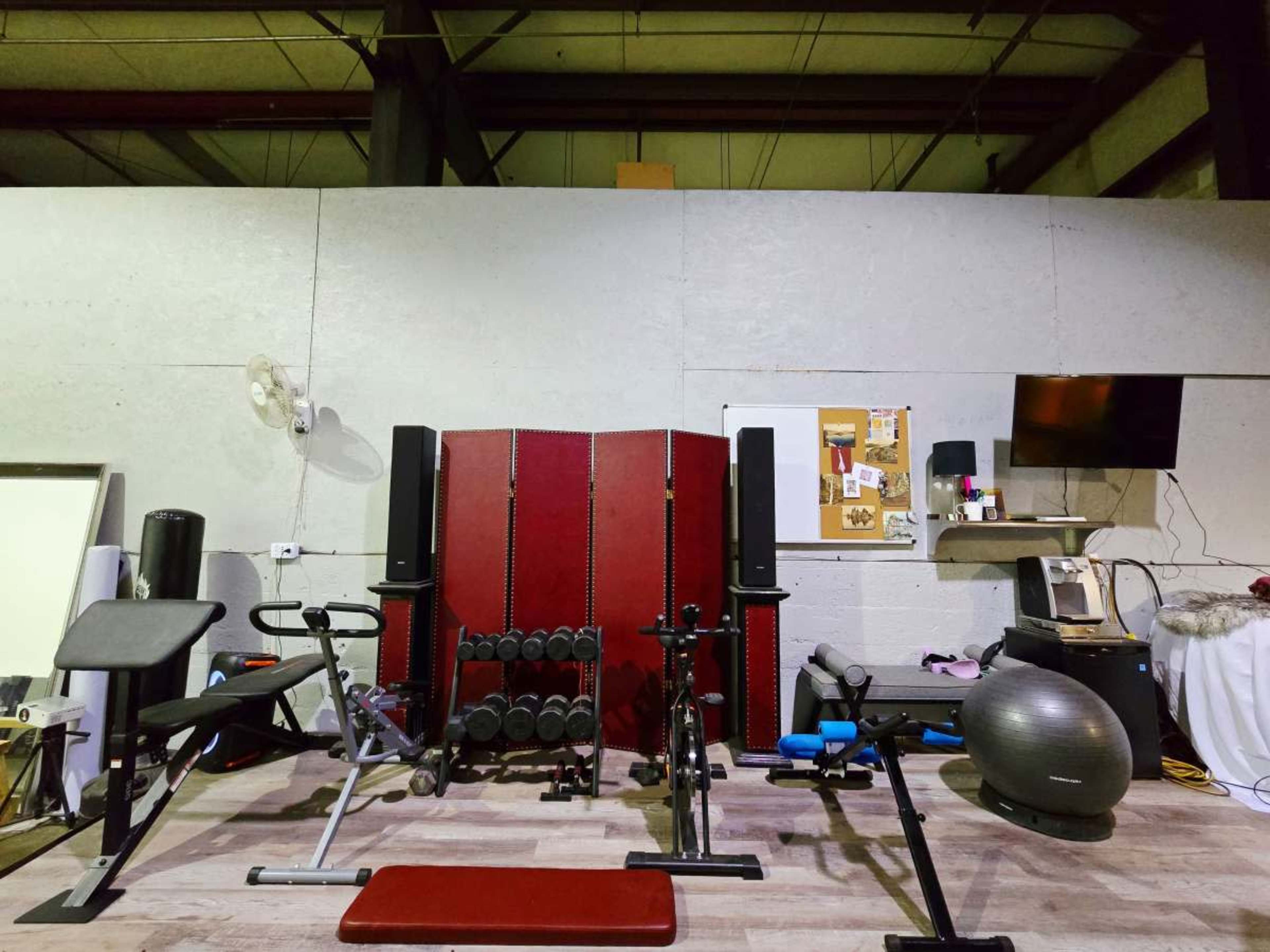 The image shows a gym space with various exercise equipments, including weights, a stationary bike, and a workout bench, against a gray wall with a bulletin board and a television.