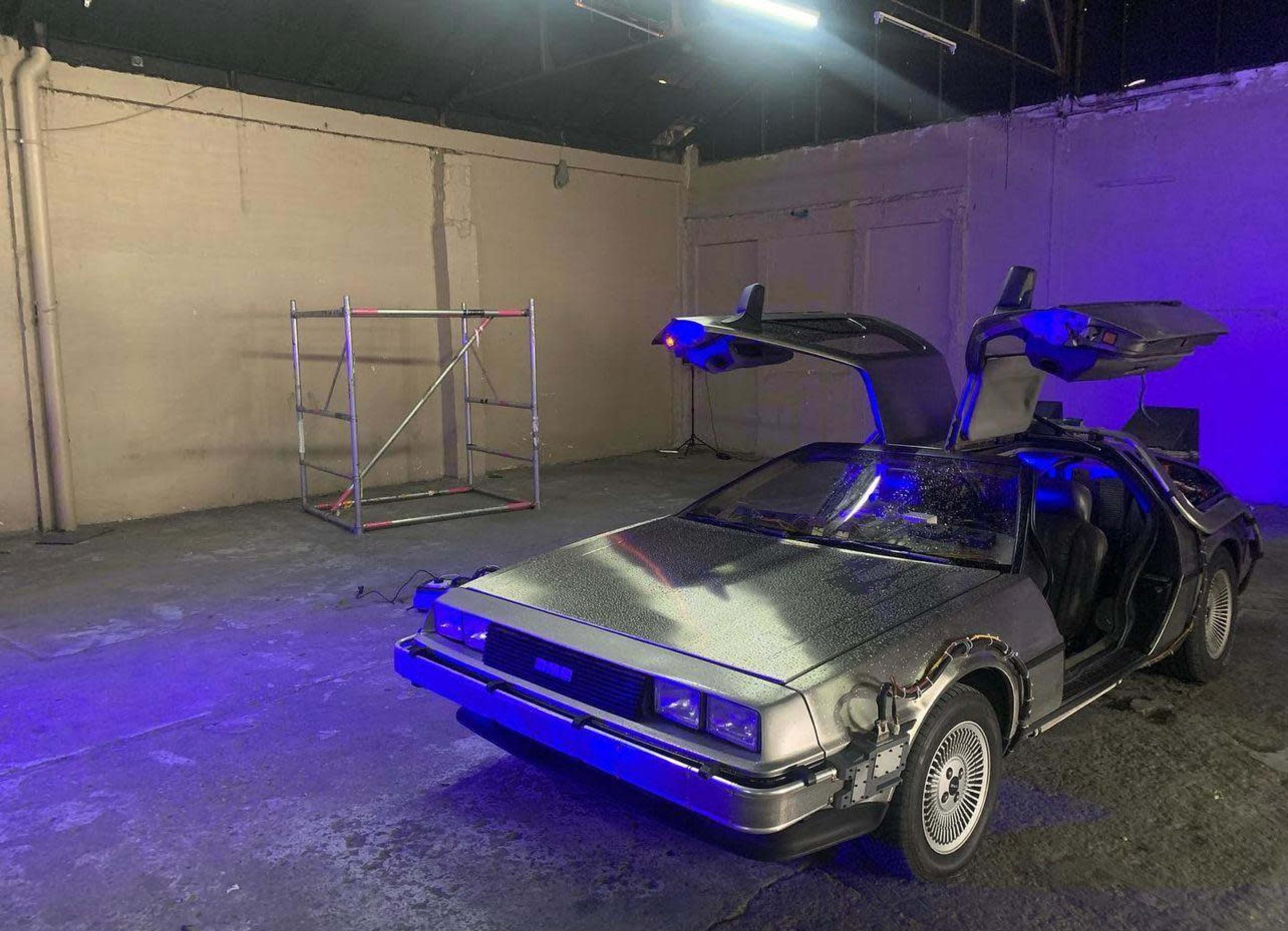 A DeLorean car with gull-wing doors is parked in an empty industrial space with a scaffold in the background and blue lighting.