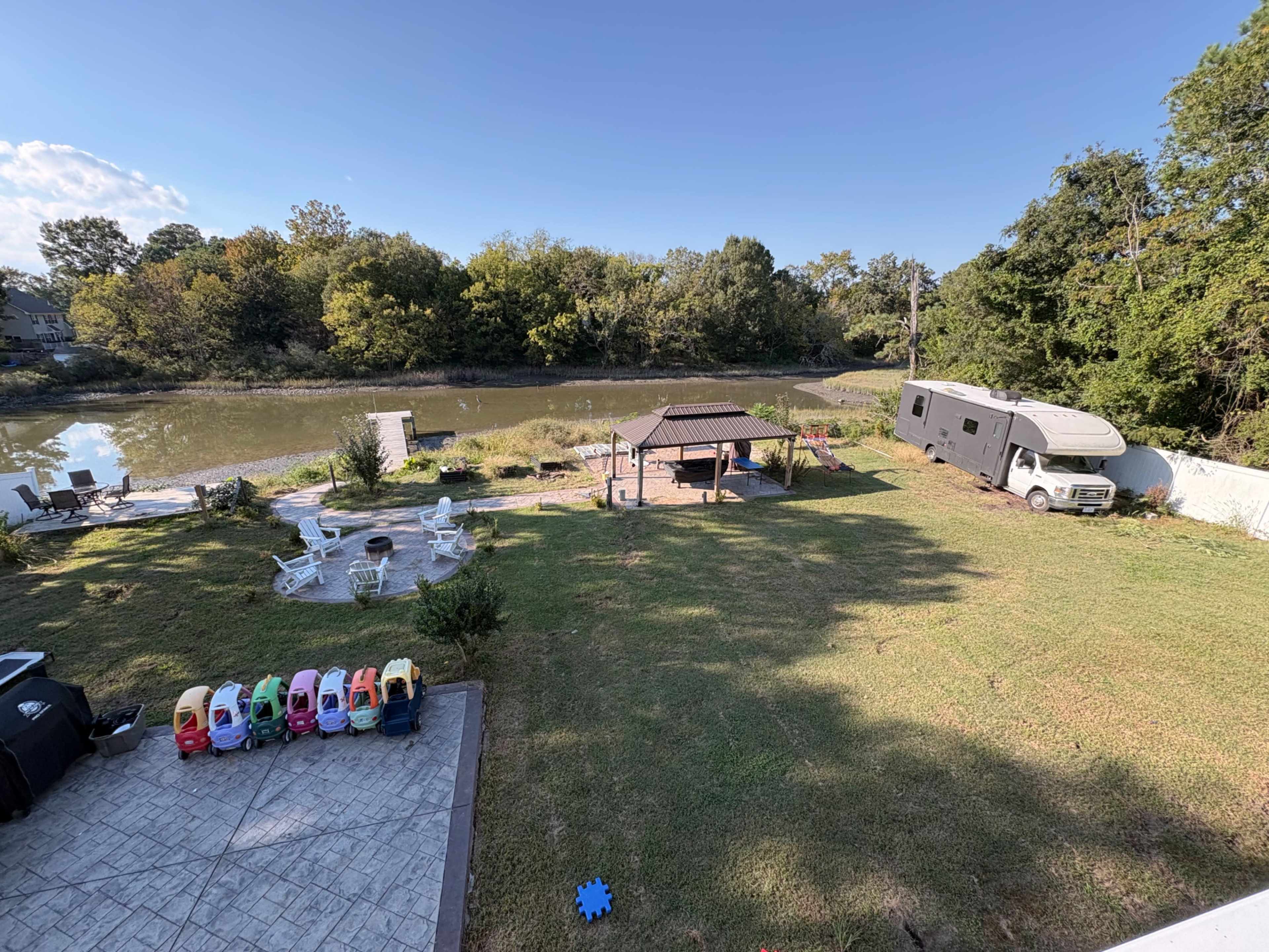 The image shows a backyard with a pond, a gazebo, an RV, a fire pit area, and several colorful children’s toy vehicles lined up.