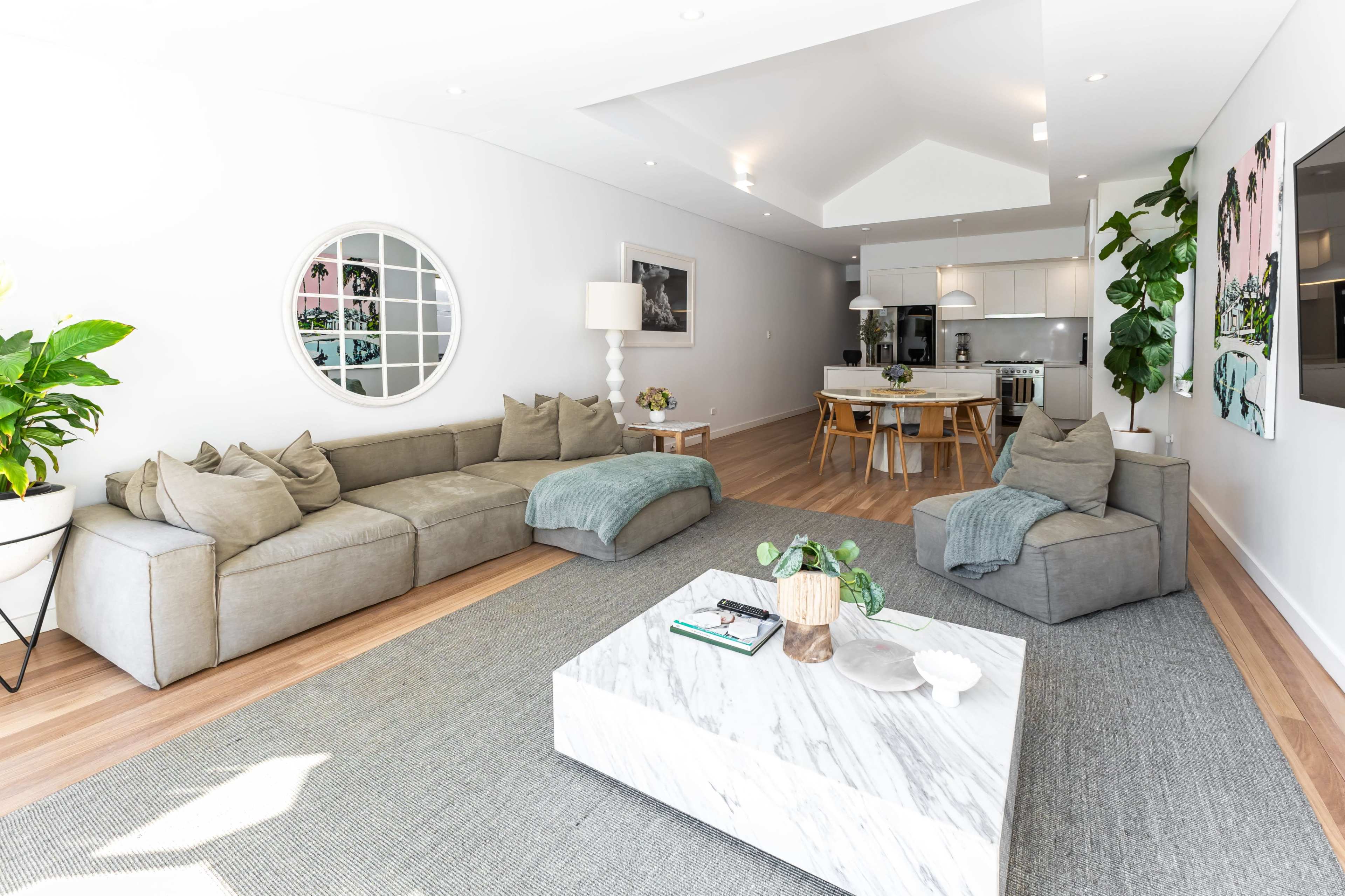 The image shows a modern living room with a gray sectional sofa, a round wall mirror, and a marble coffee table, set against a backdrop of light-colored walls and wooden flooring.