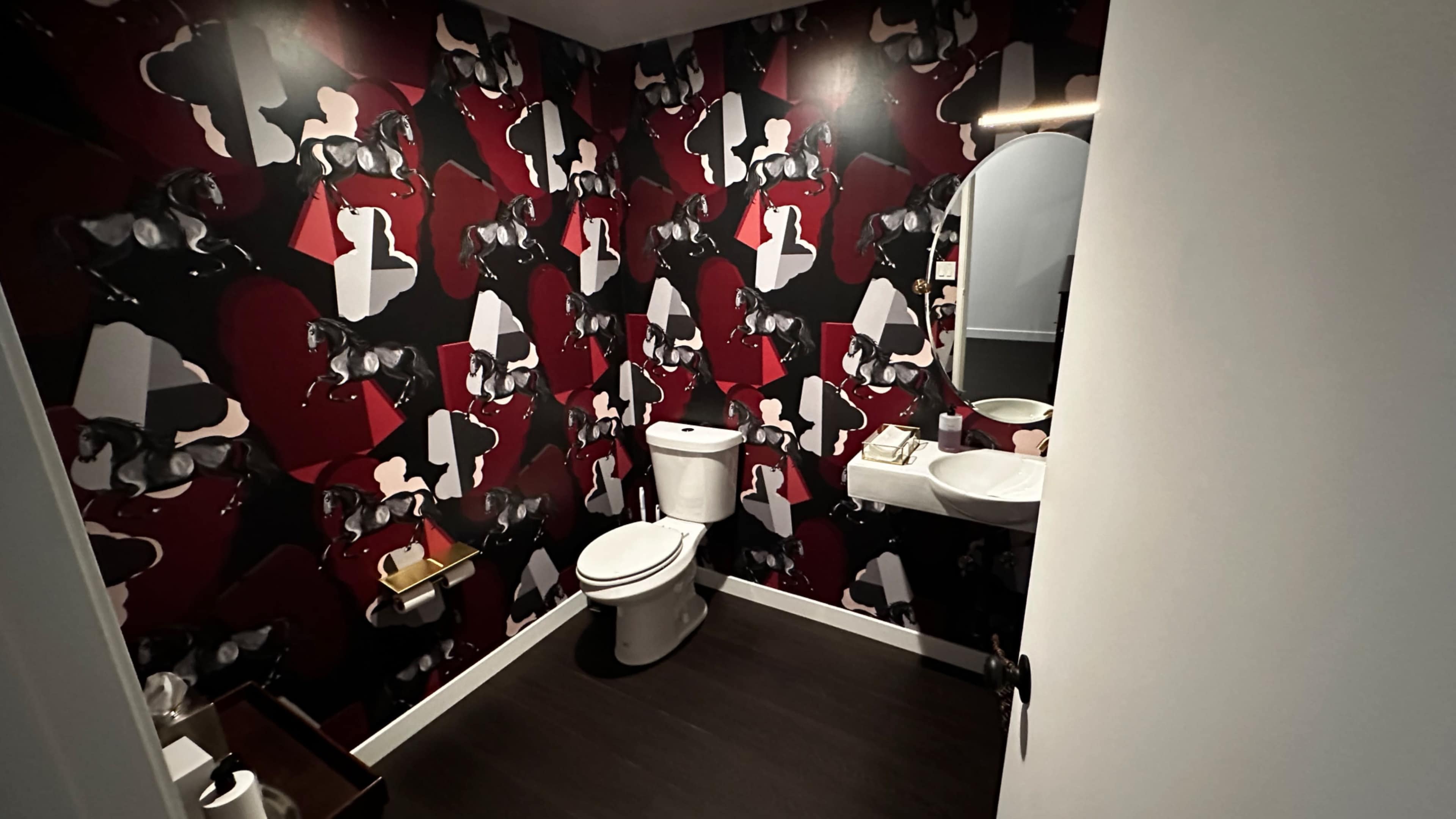 A small bathroom features patterned red and black wallpaper, a white toilet, and a sink with a round mirror.