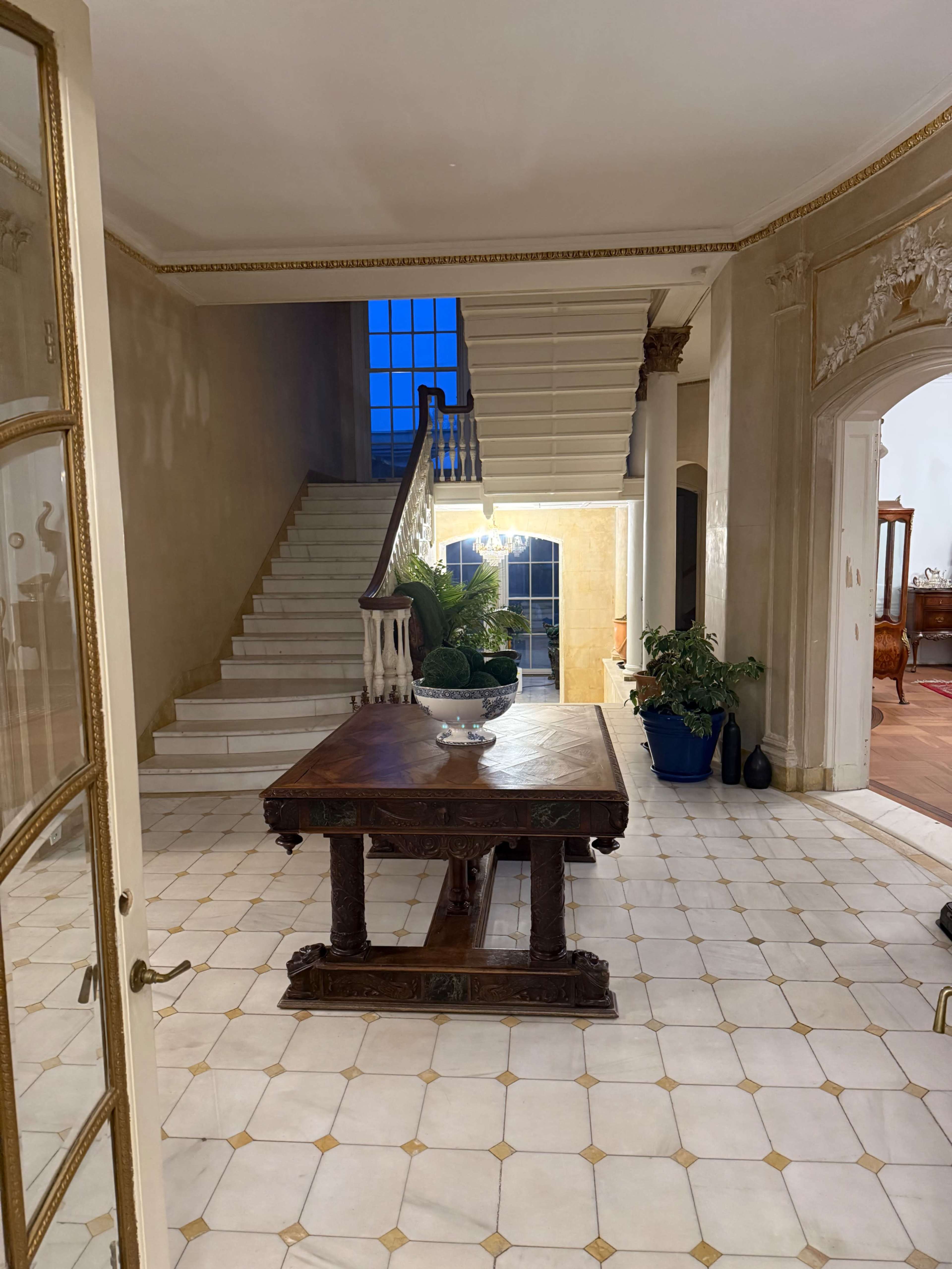 The image shows a spacious entryway featuring a large wooden table, a staircase in the background, and potted plants on either side.