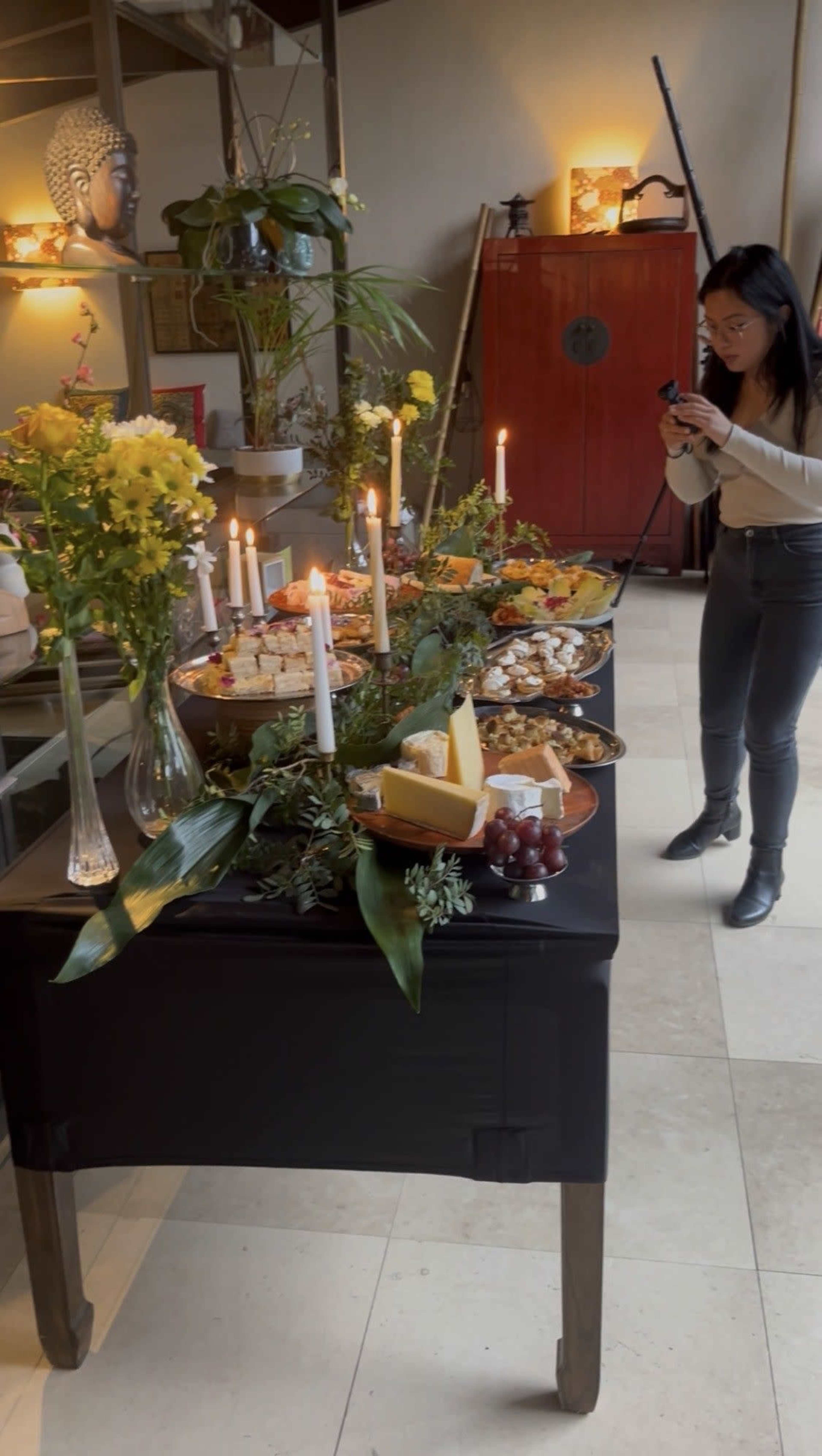 A woman takes a photo of a decorated table featuring a variety of cheeses, fruits, and candles in a stylish indoor setting.