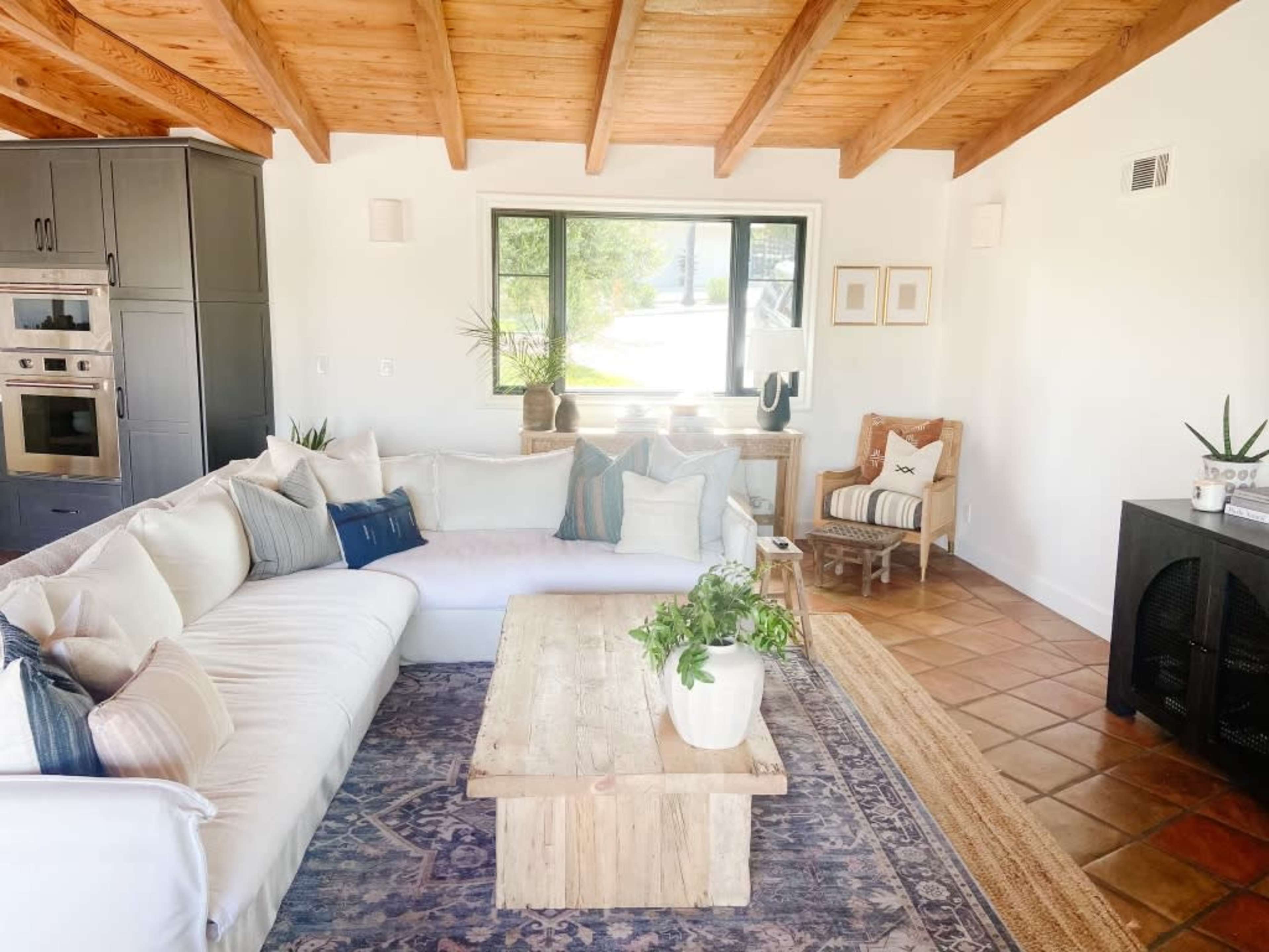 The image shows a cozy living room with a large sectional sofa, a wooden coffee table, and a mix of plants and decorative elements against a backdrop of exposed wood beams and large windows.
