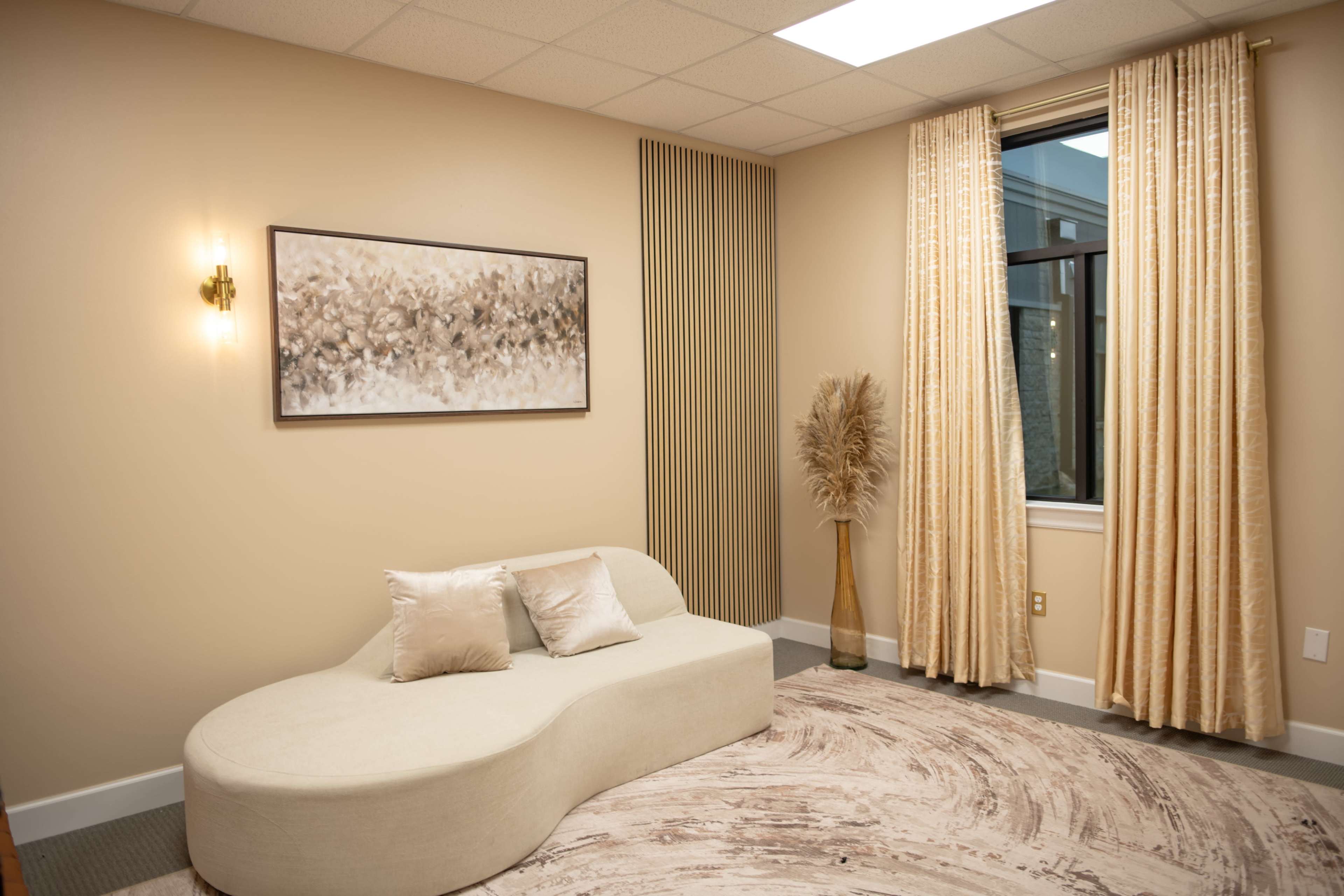 The image shows a softly curved white lounge sofa positioned against a light-colored wall, complemented by elegant curtains and a decorative plant in a vase.