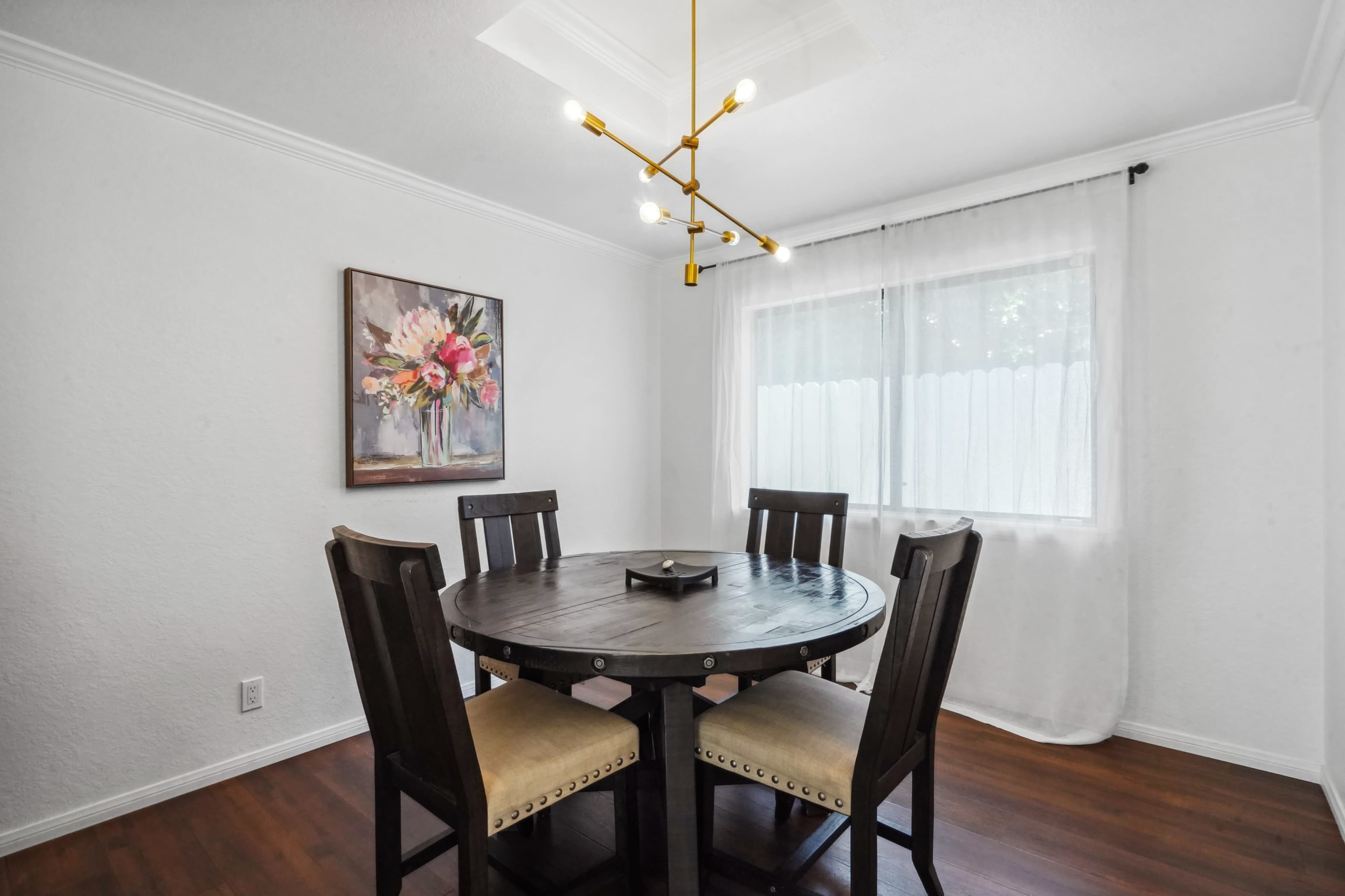 A round wooden dining table with four chairs is placed in a well-lit room that features a floral painting and a window covered with sheer curtains.