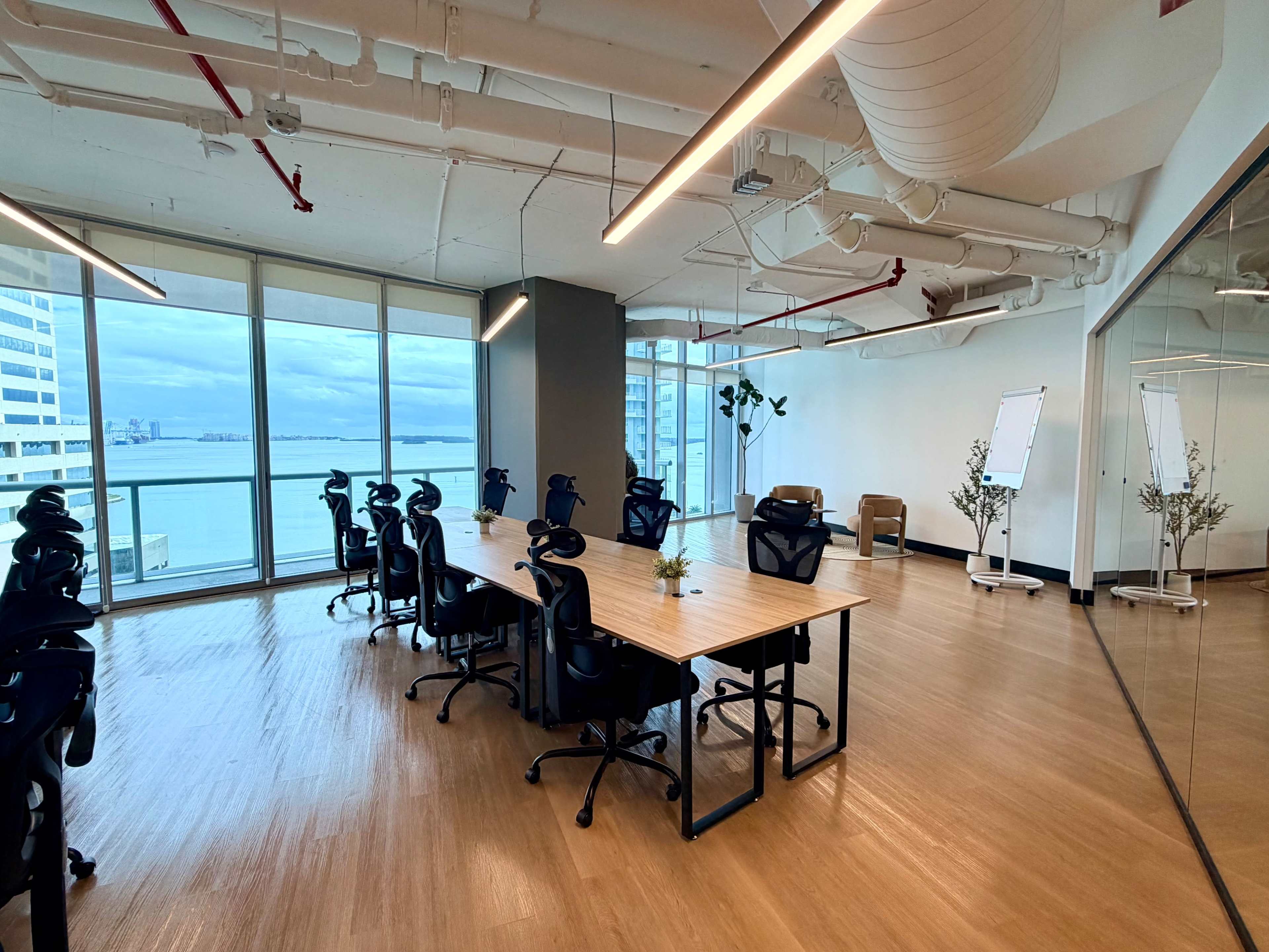 A modern, empty conference room features a large table surrounded by black office chairs, with windows that offer a view of the waterfront and a few plants decorating the space.