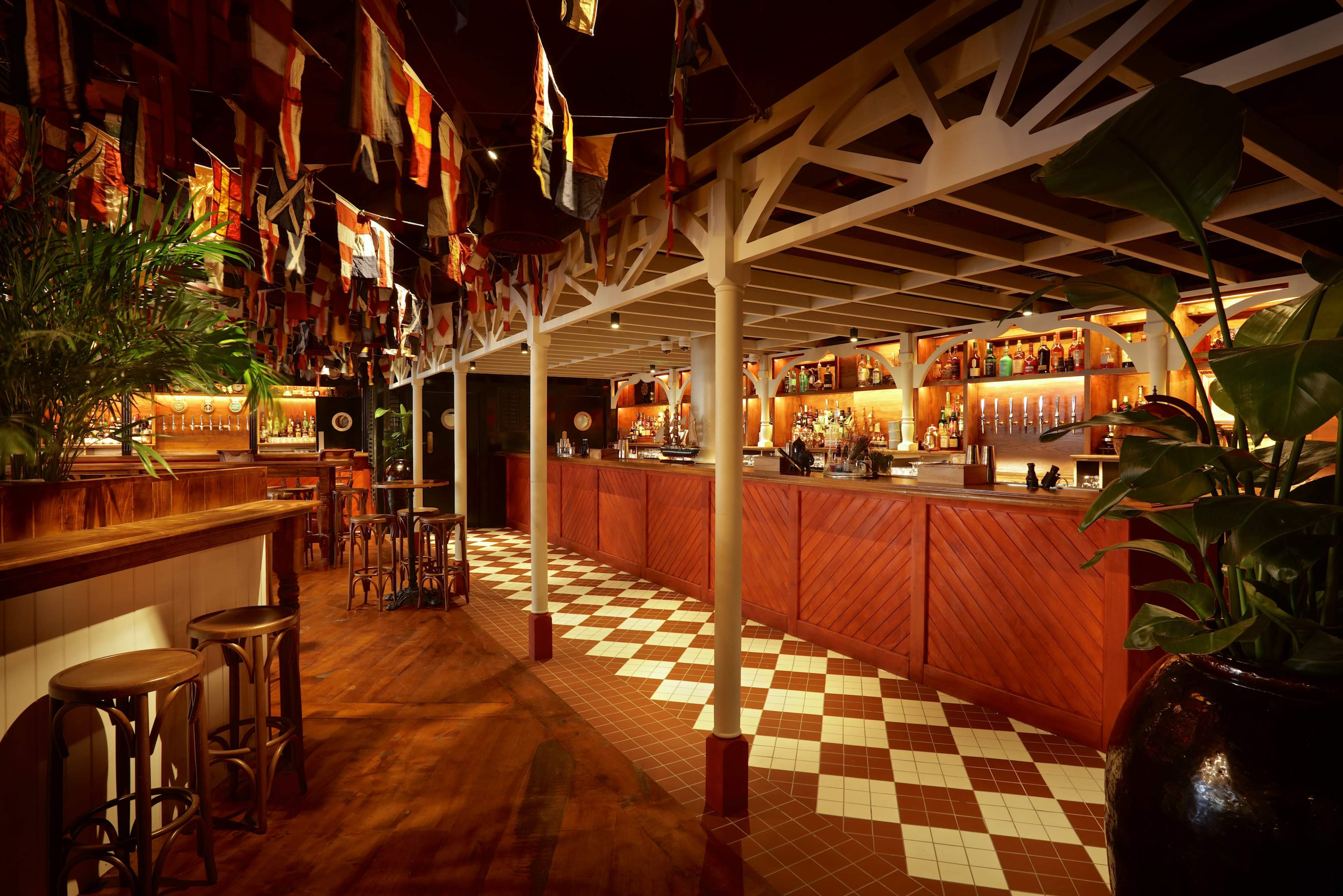 The image shows a warmly lit bar interior with wooden furnishings, a long counter stocked with bottles, and colorful flags hanging from the ceiling.