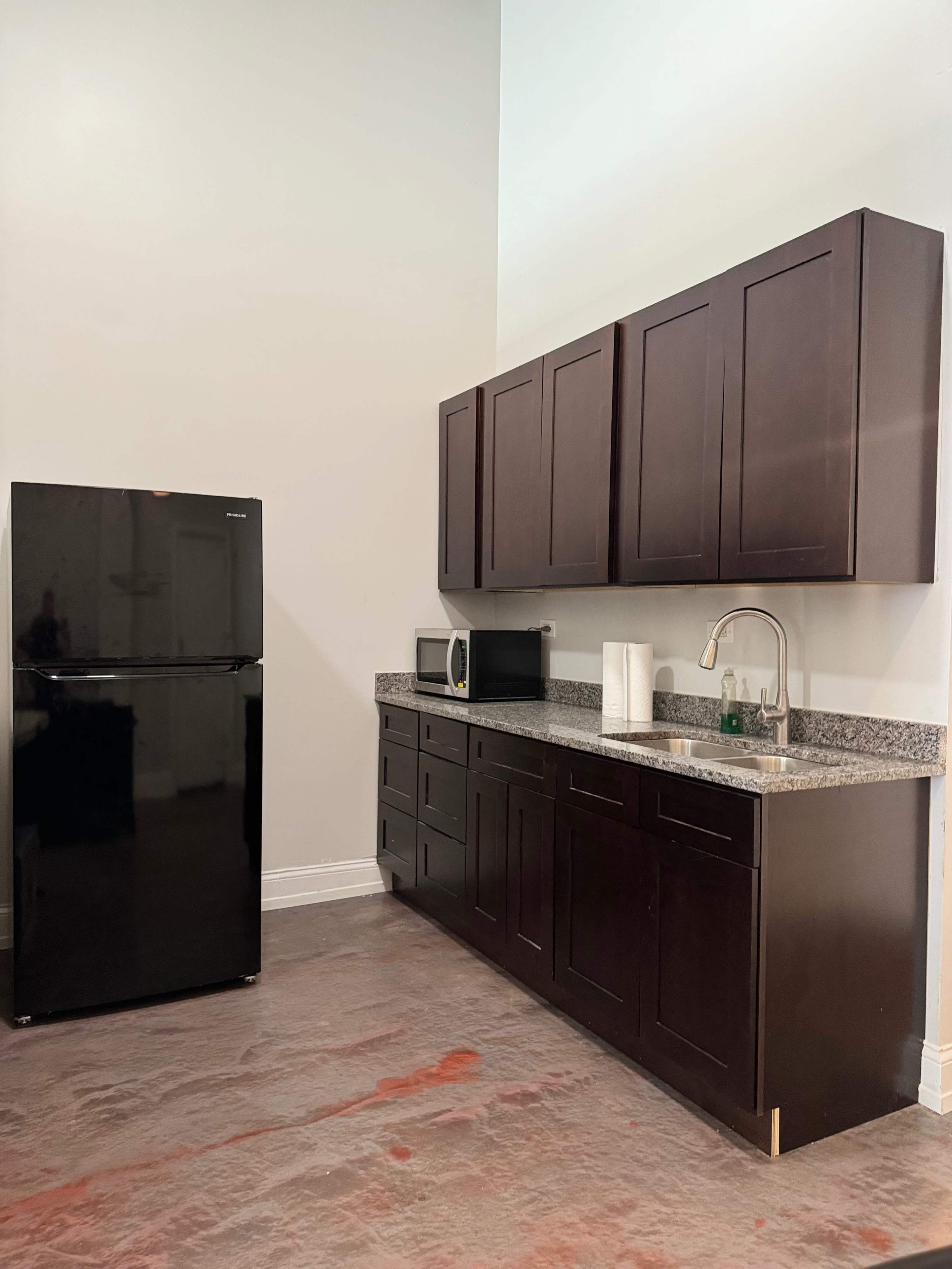 A kitchen with dark cabinetry and a black refrigerator, set against a light-colored wall and a floor covered in a pinkish material.