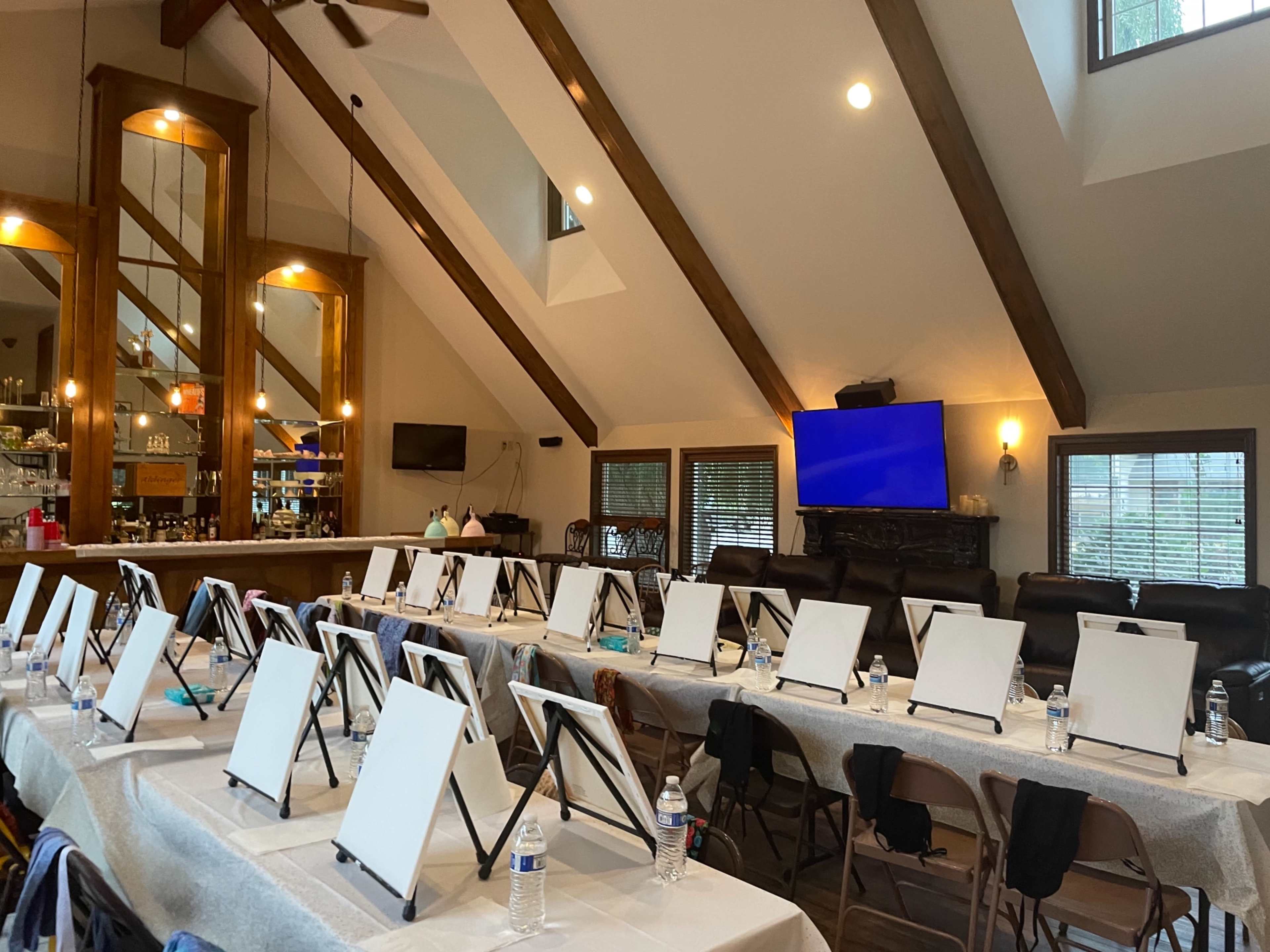 An indoor space set up for a painting class, featuring multiple empty easels arranged neatly on tables with water bottles and a bar area visible in the background.