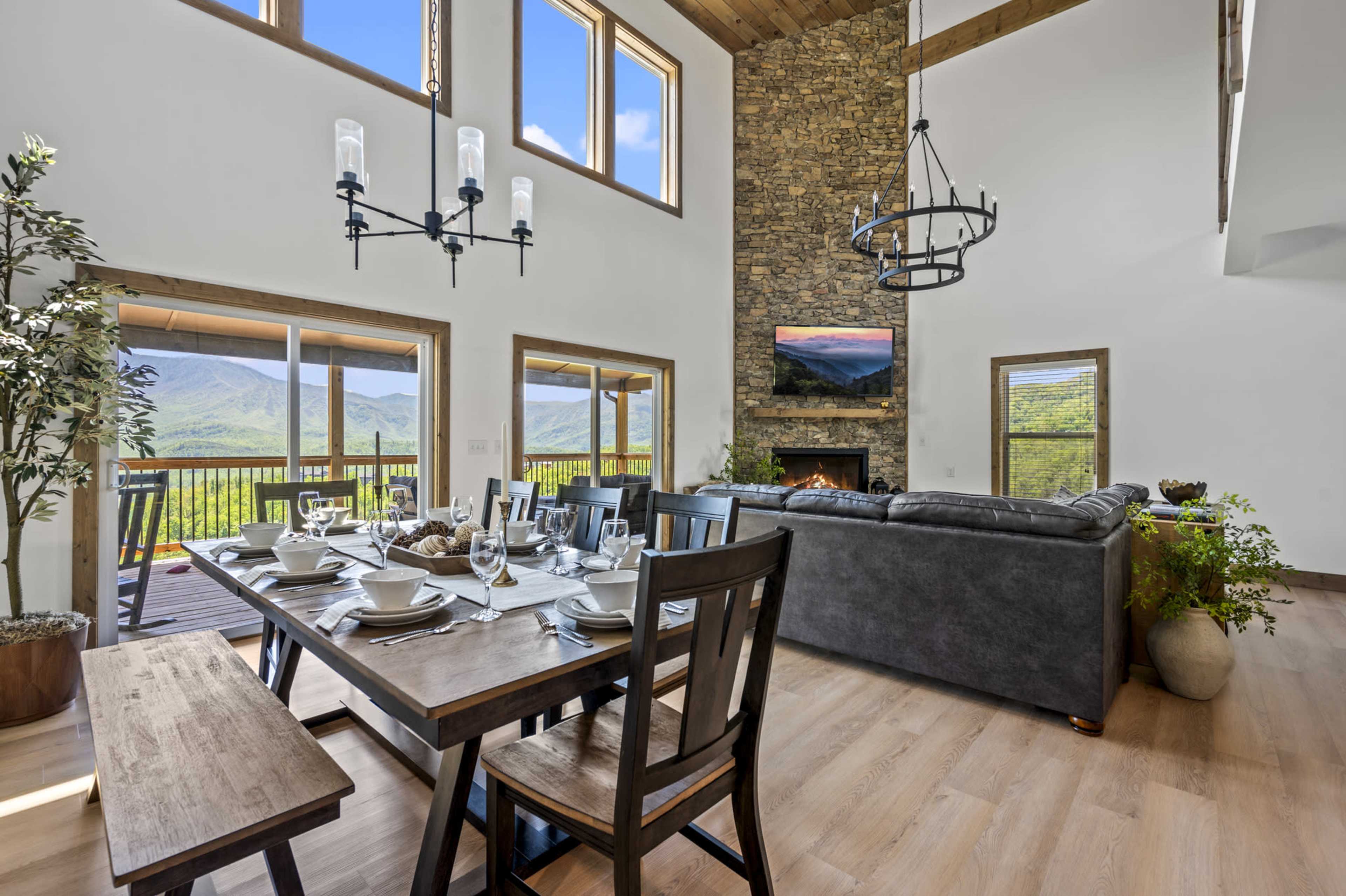 A dining area with a large wooden table set for a meal, adjacent to a living space featuring a stone fireplace and large windows that overlook a mountain view.