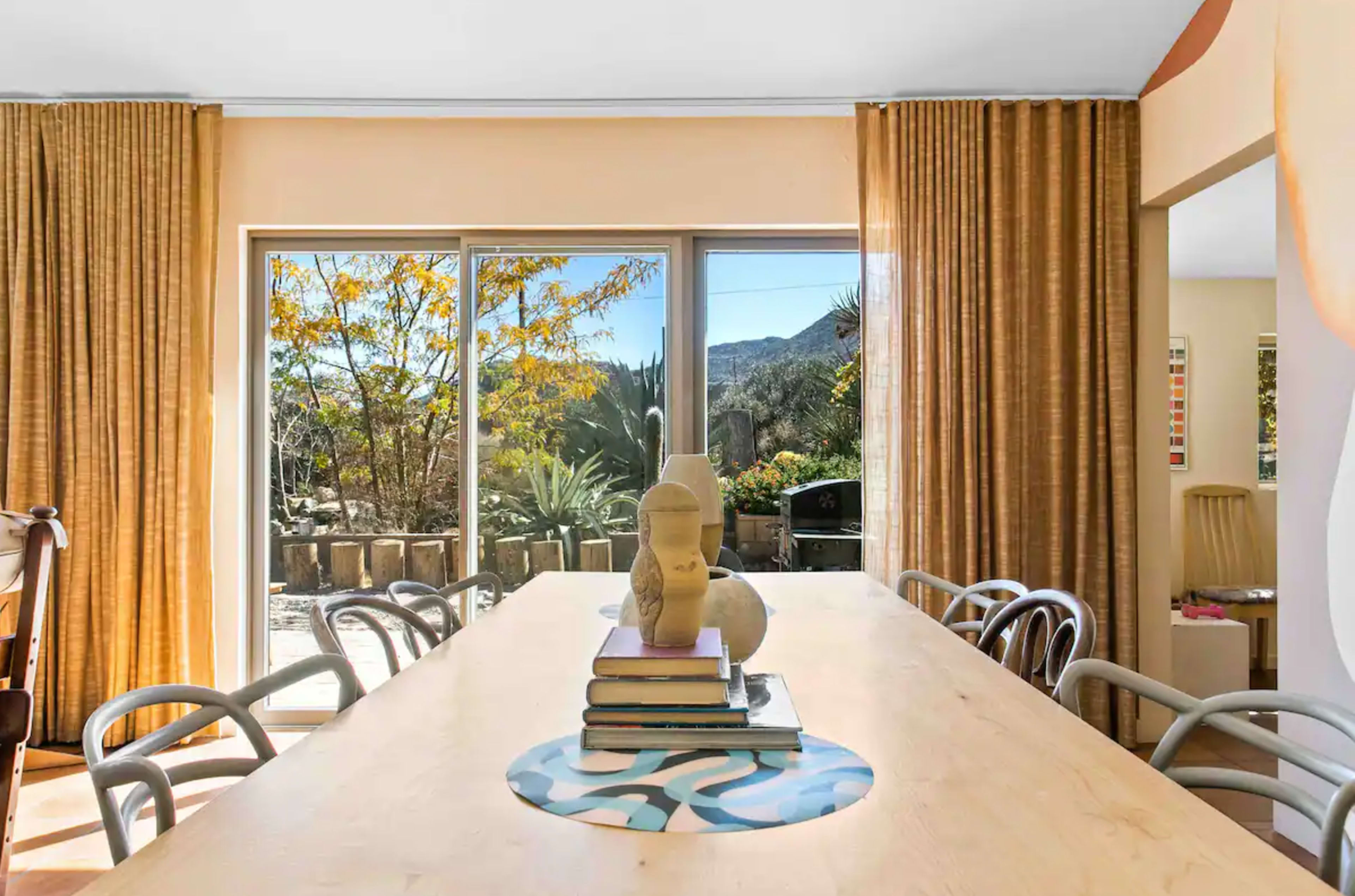 A dining table with books and a sculpture is positioned in front of large windows revealing a view of an outdoor area with plants and mountains.