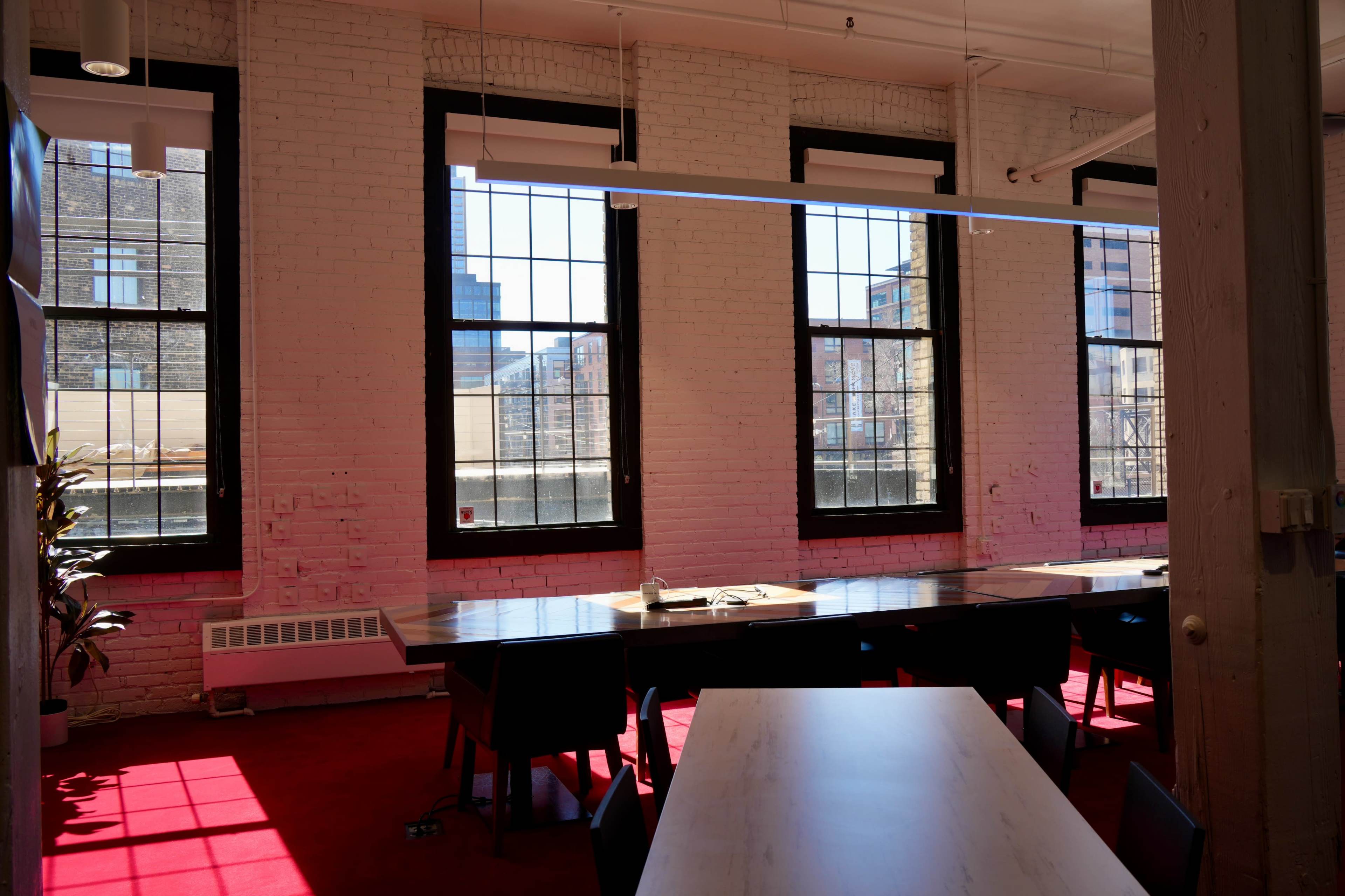 The image shows a bright office space with large windows, red carpet, and a long table surrounded by chairs.