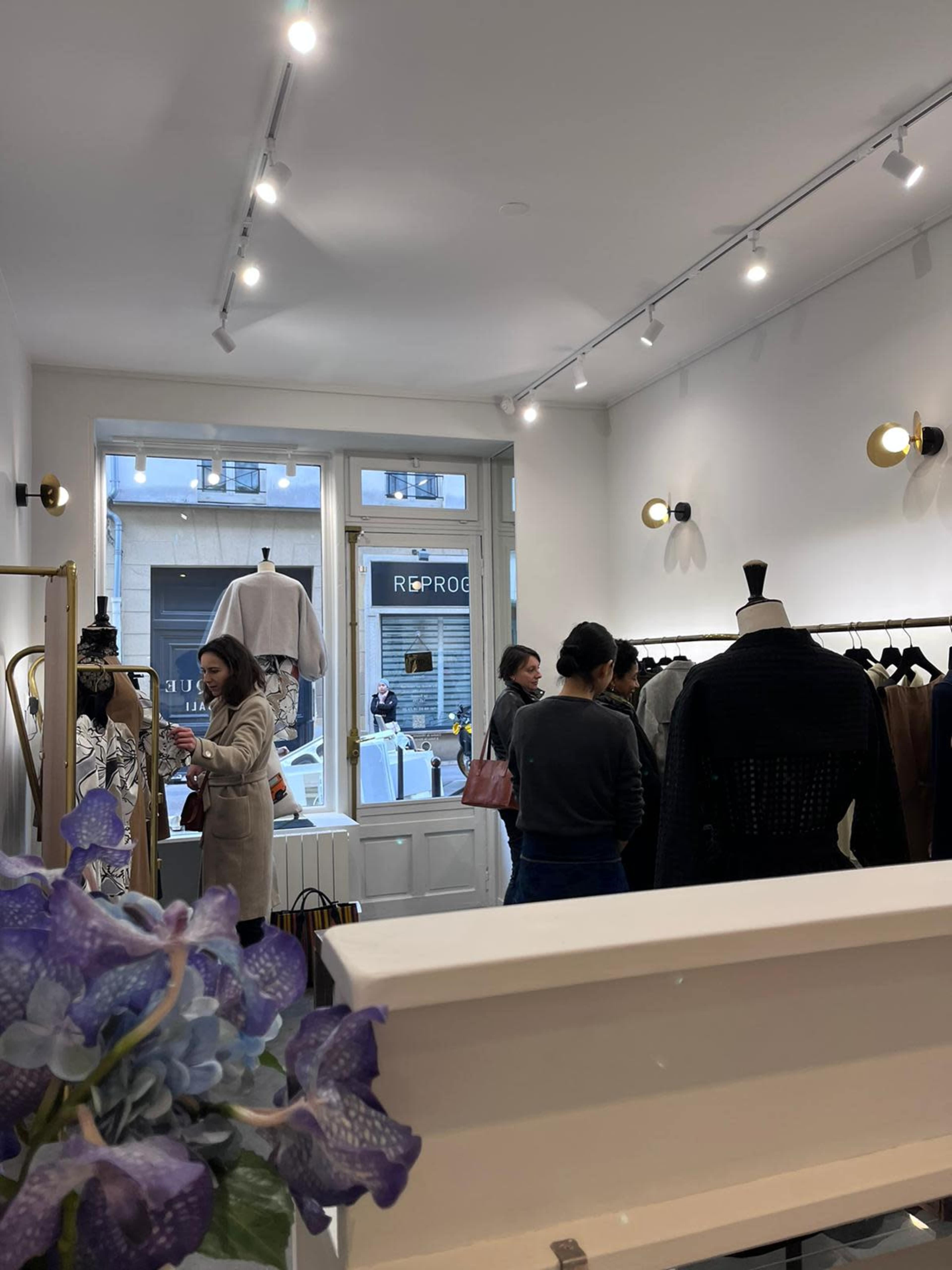 A group of shoppers browse clothing in a boutique with large windows and clothing racks.