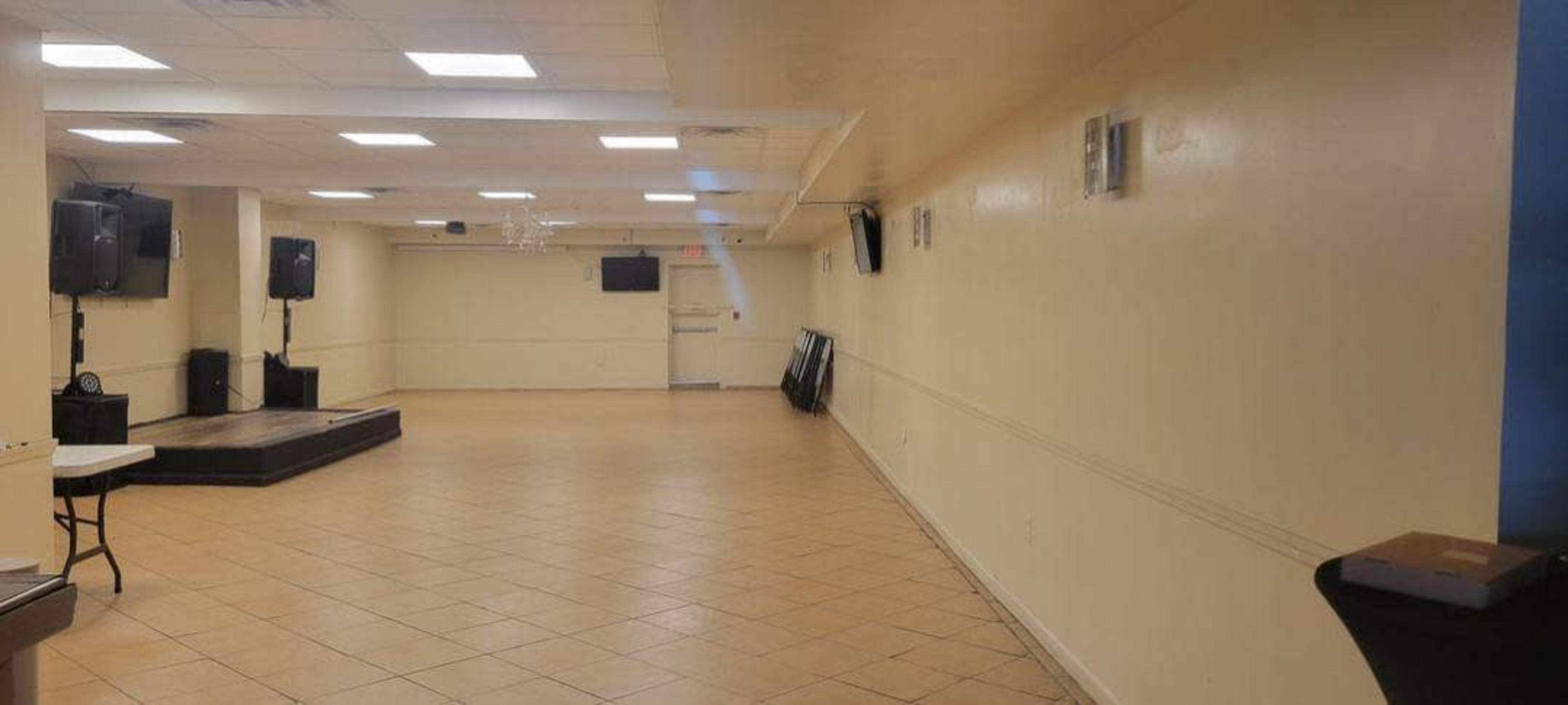 An empty event space with beige walls, tiled flooring, and two large speakers positioned on stands at opposite ends.