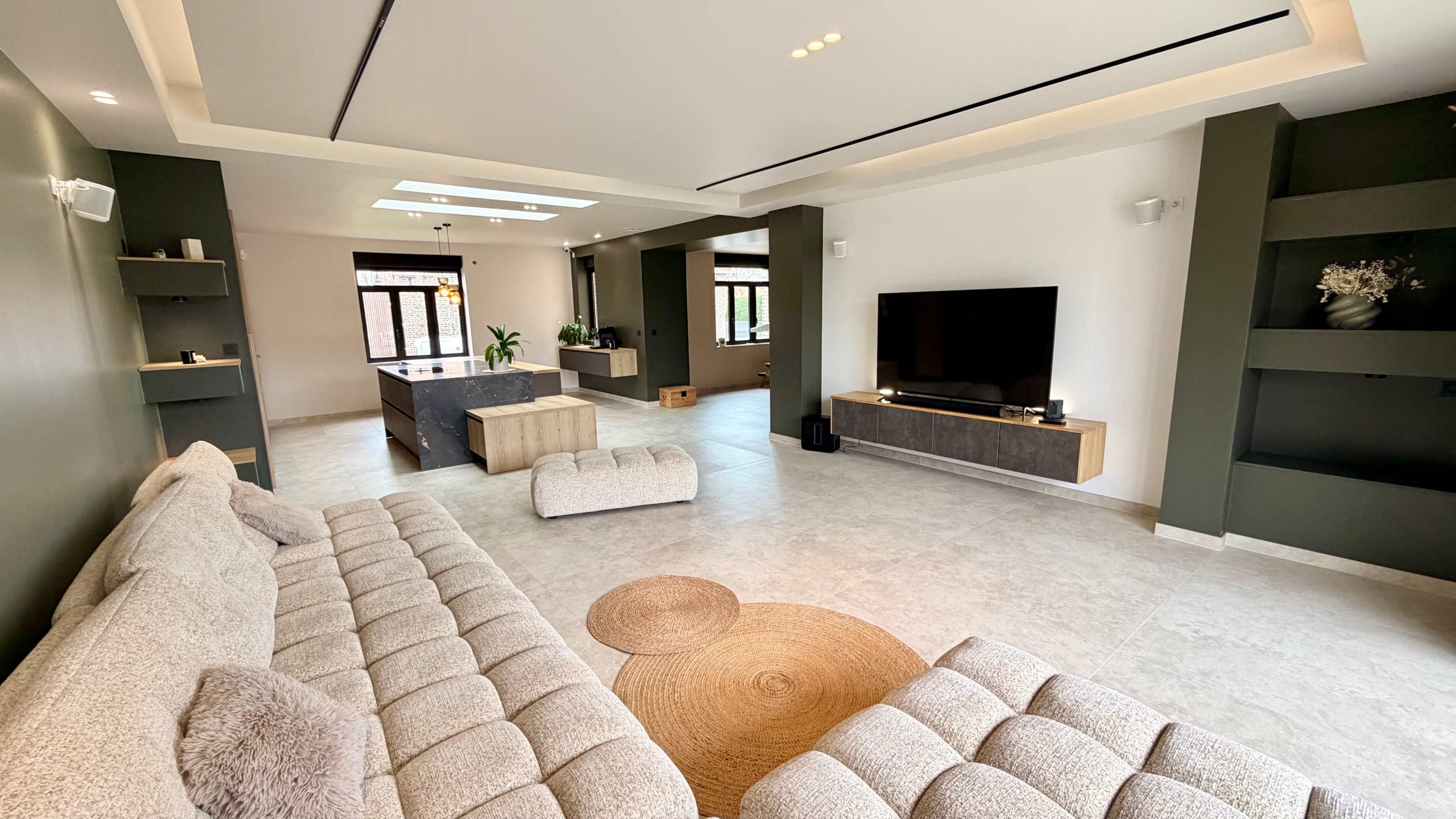 The image depicts a spacious living room featuring a large, light-colored sectional sofa, a circular area rug, and a mounted television with a wooden media console against a neutral wall.