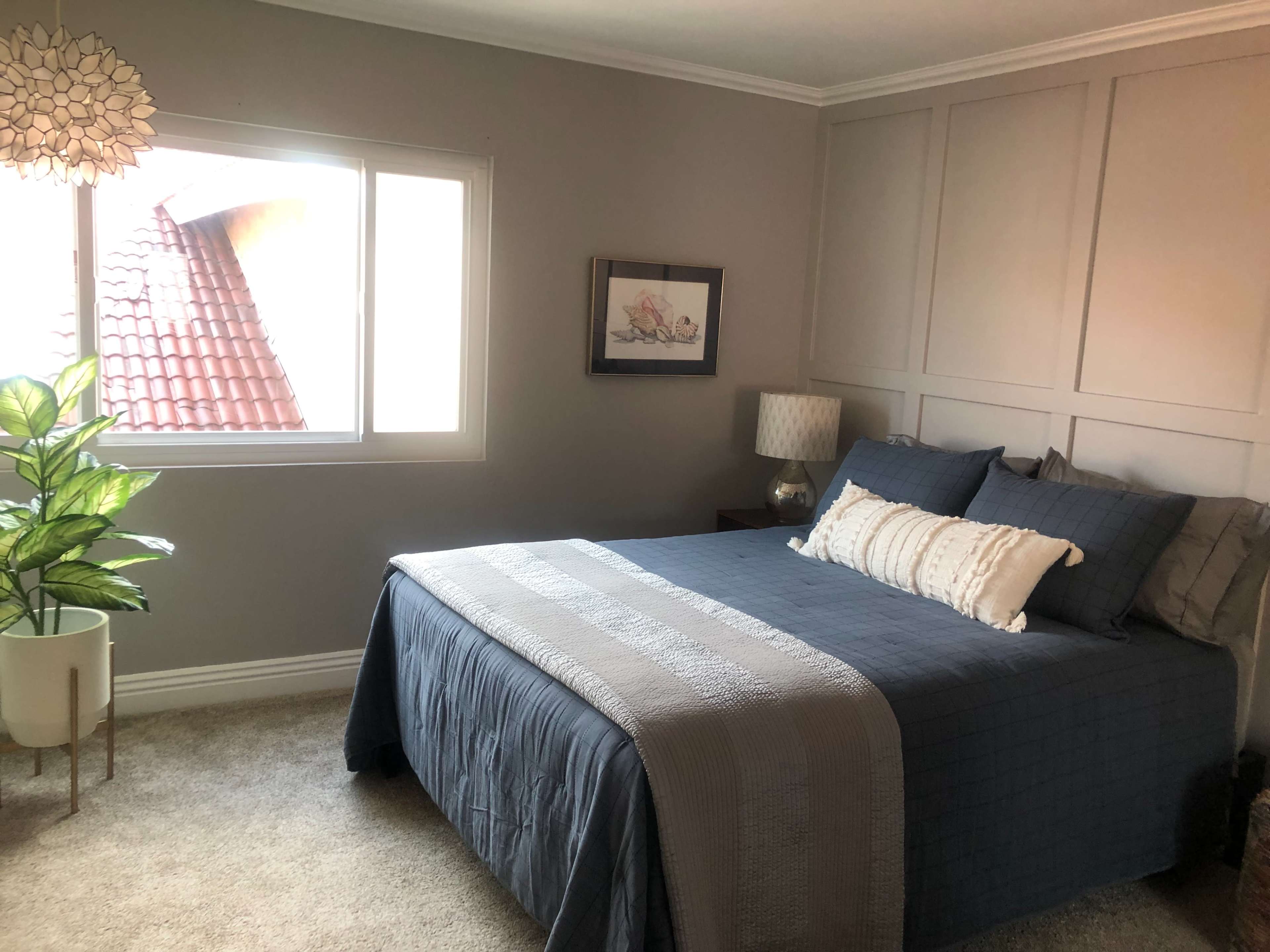 A neatly arranged bedroom features a bed with blue bedding, a window allowing natural light, and a potted plant in the corner.