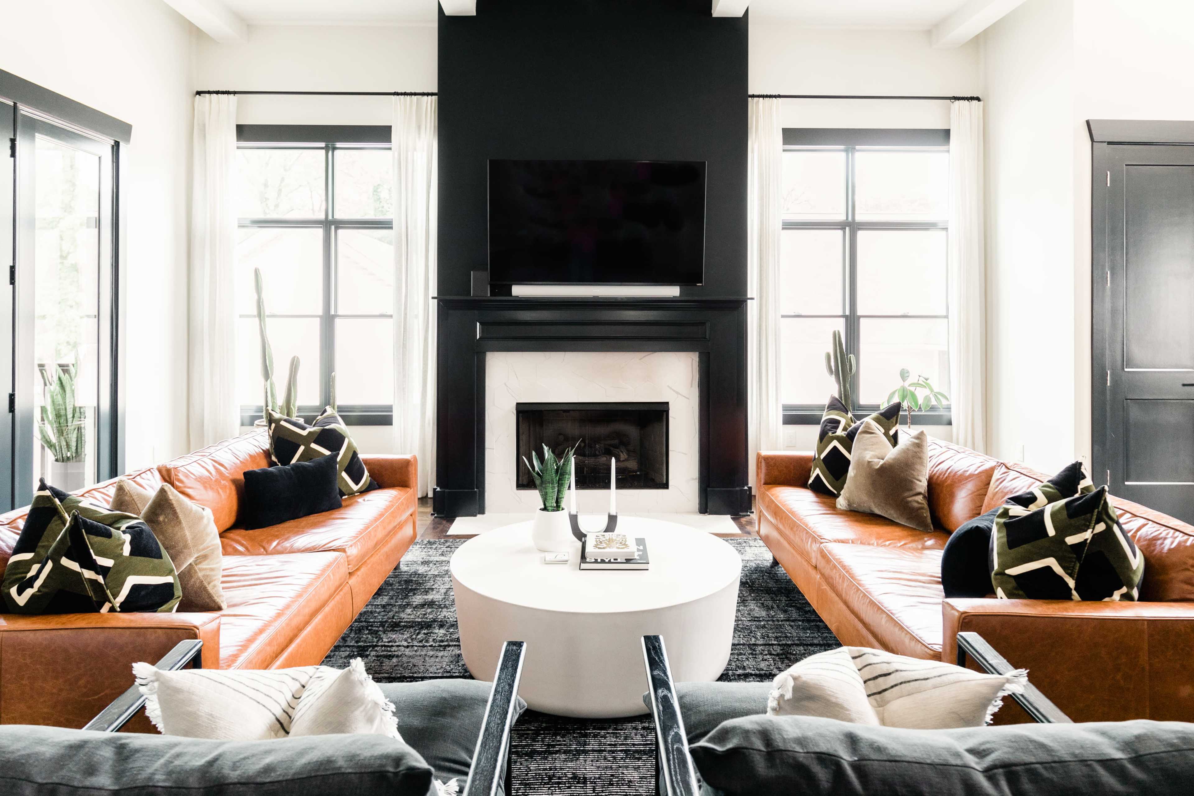 A modern living room features two leather sofas, a circular coffee table, a large fireplace with a TV above it, and large windows with plants on either side.