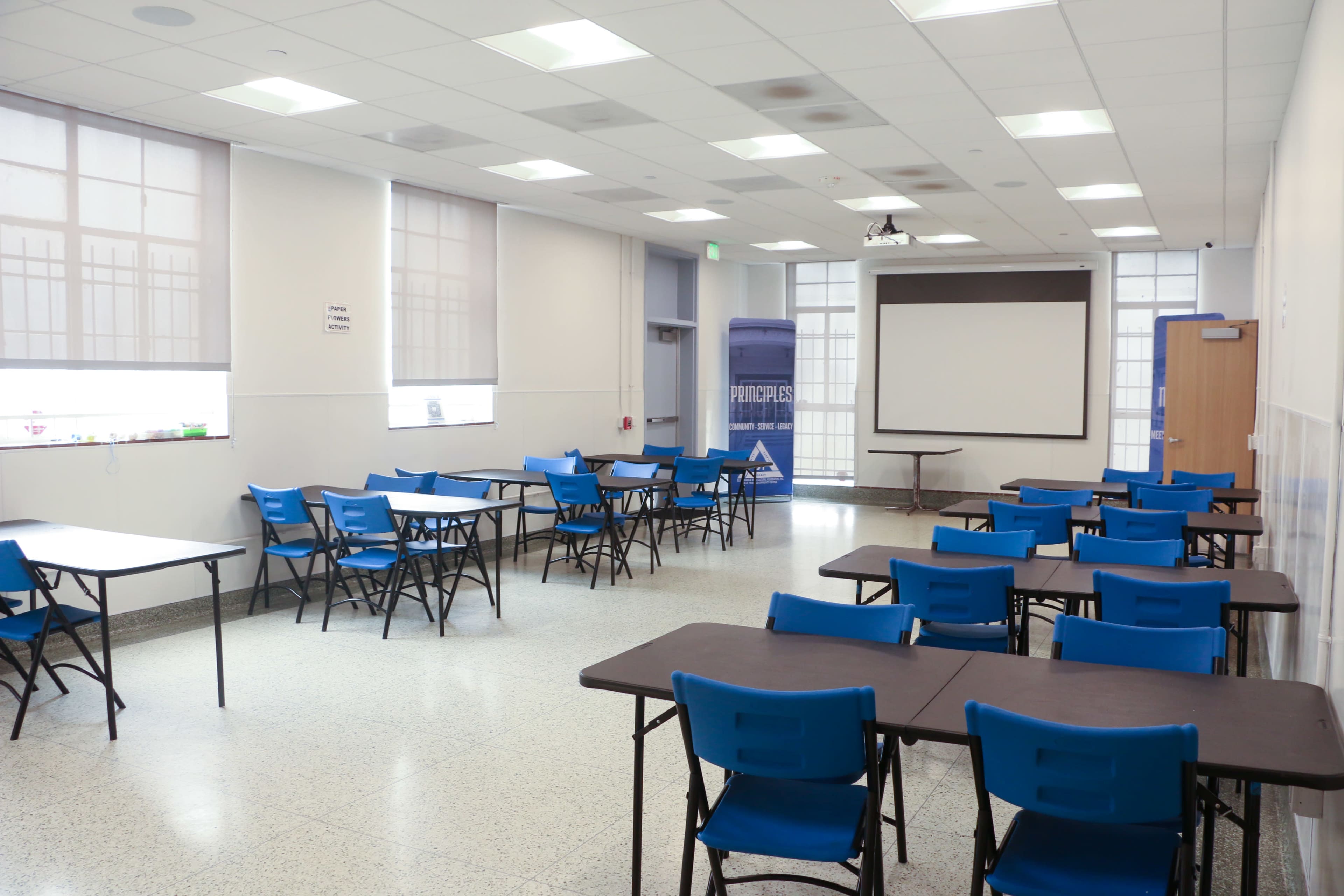 The image shows an empty classroom with blue chairs and tables arranged in rows, featuring large windows with shades and a screen mounted on the wall.