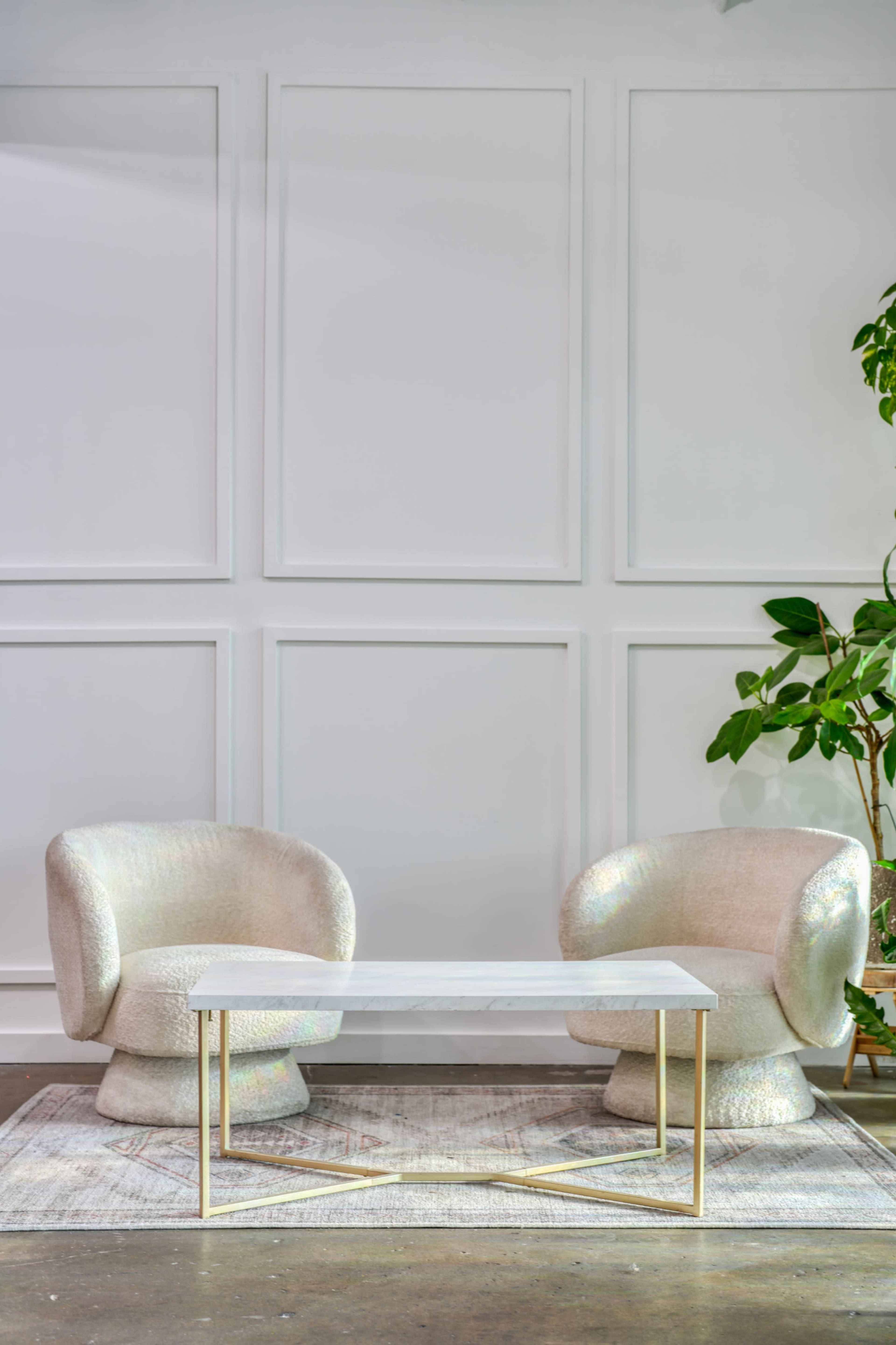 The image shows a minimalist seating area featuring two plush, curved chairs and a marble-top coffee table, positioned against a plain white wall with a decorative panel design and a potted green plant nearby.