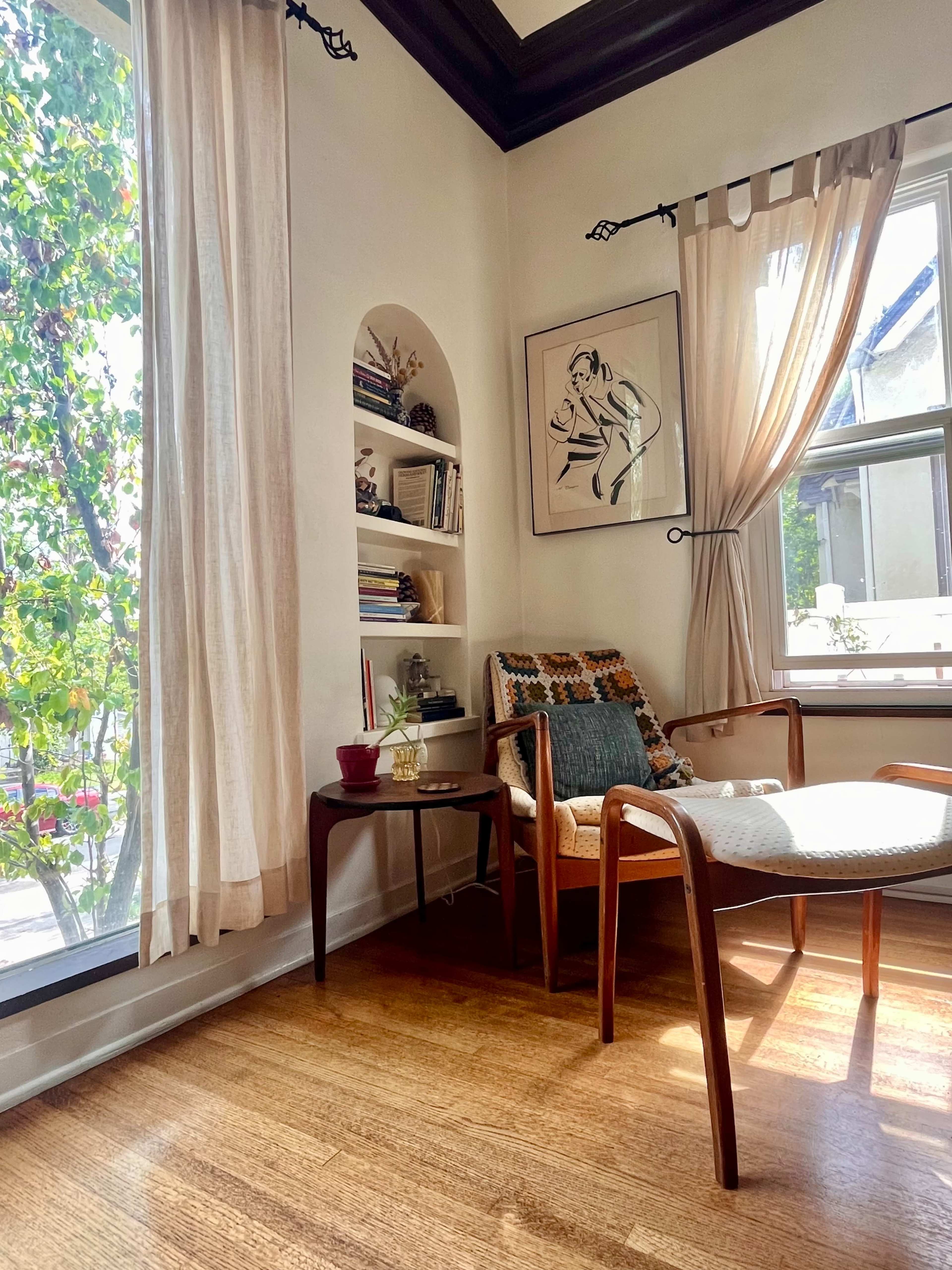 A cozy corner of a room features a wooden chair, a small side table, and shelves filled with books, illuminated by natural light streaming through large windows.