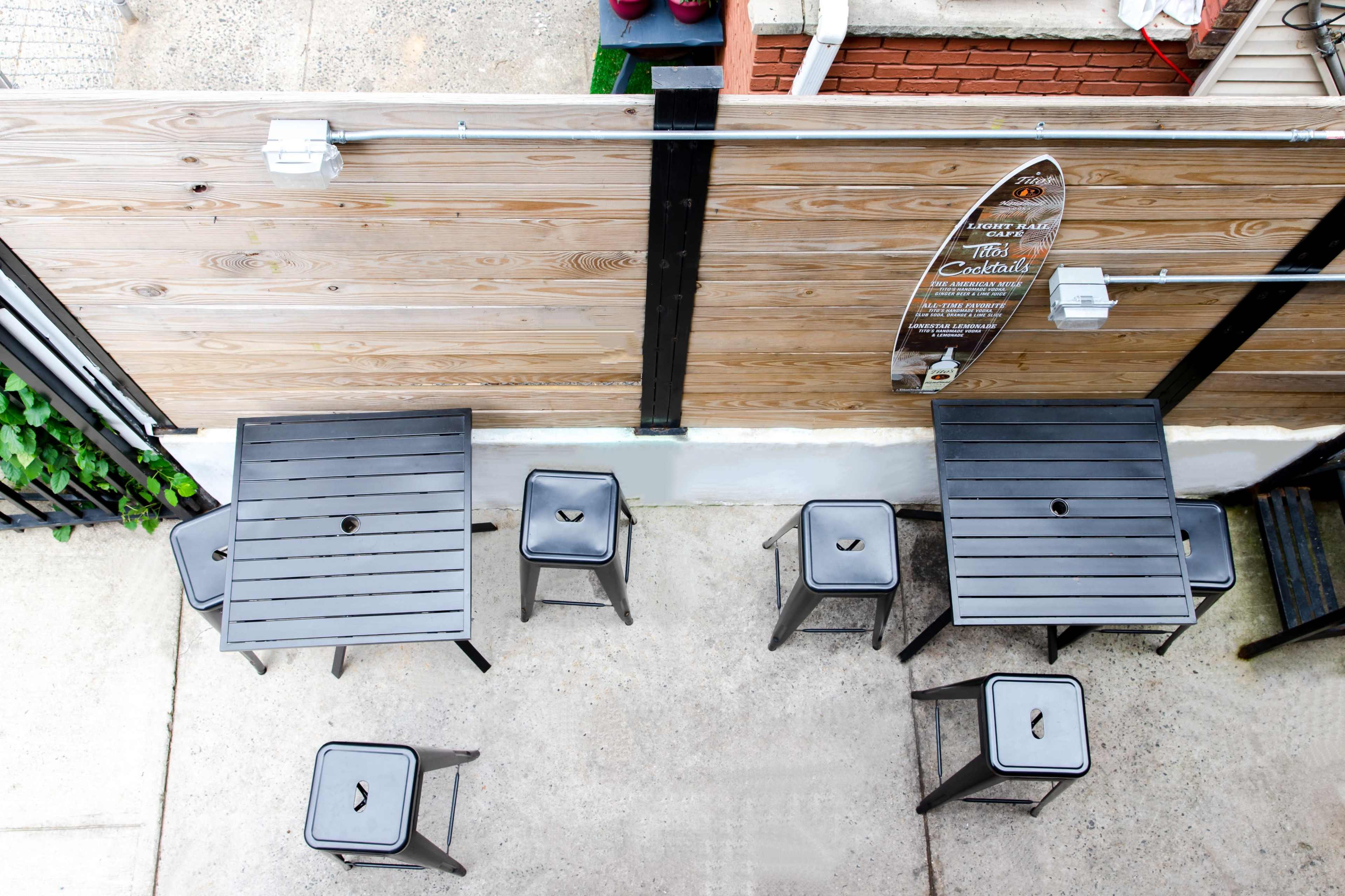 A collection of black metal tables and stools is arranged on a concrete surface against a wooden fence, with a surfboard mounted on the wall.