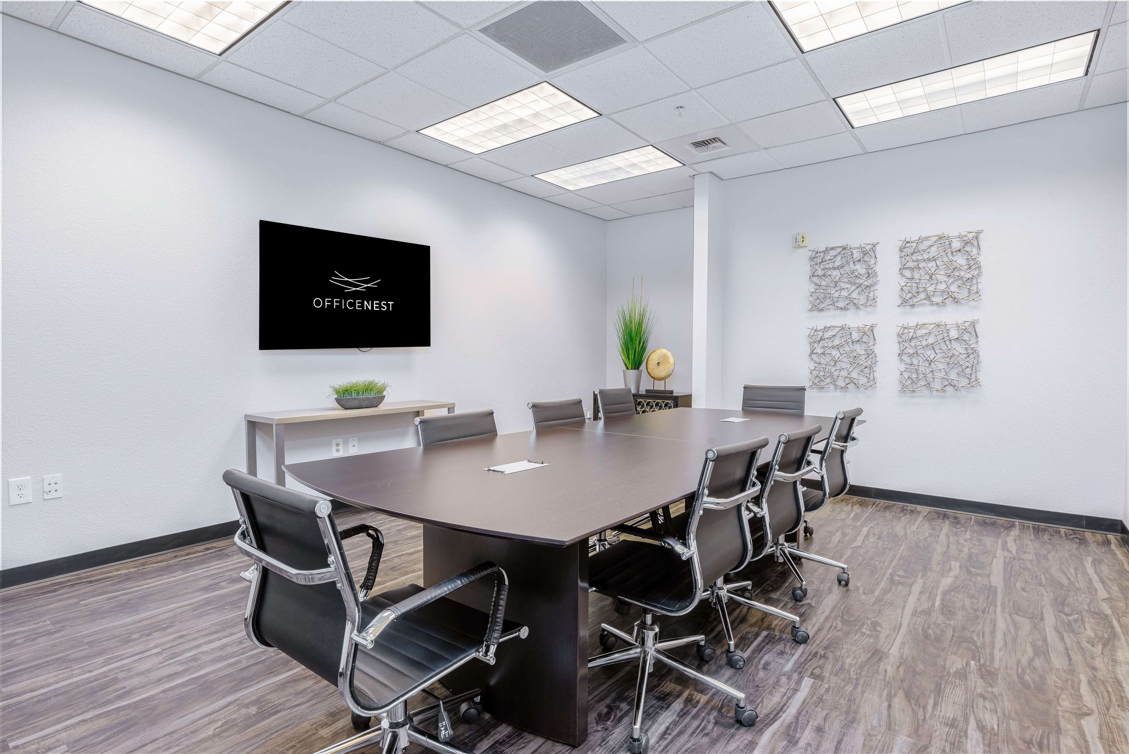 A modern conference room features a long table surrounded by ergonomic chairs, with a wall-mounted TV and decorative wall art.