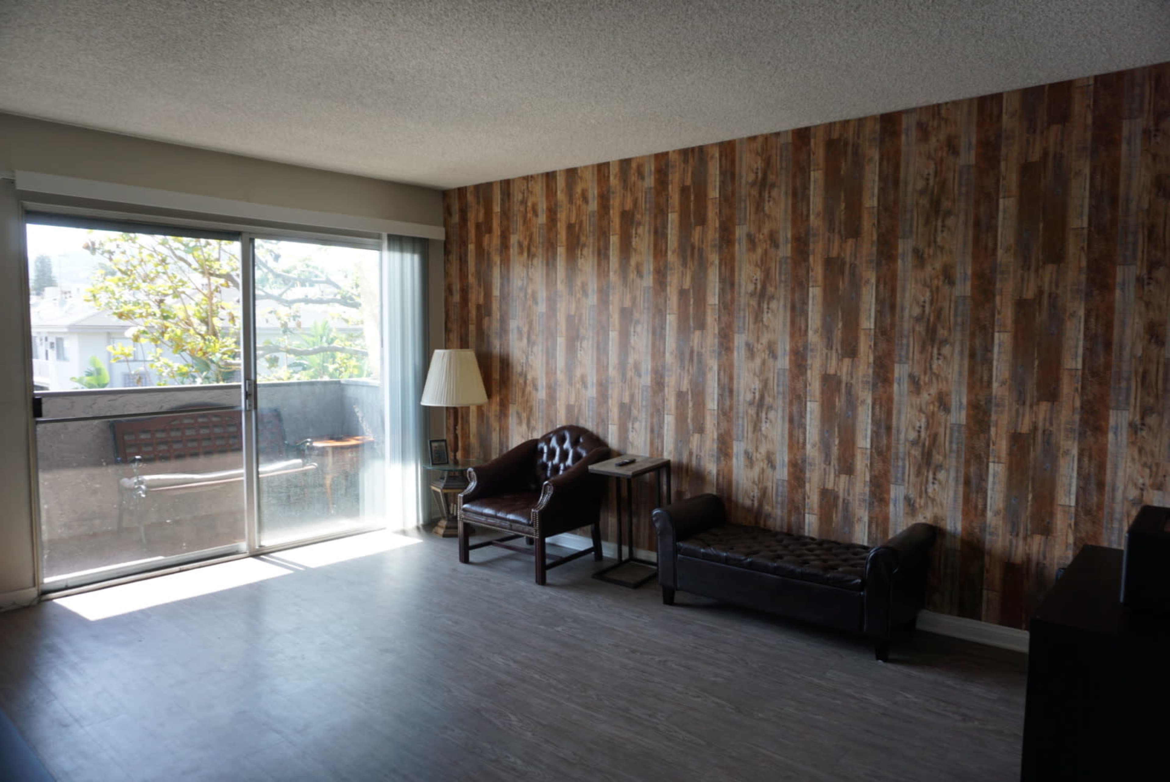 A living room with wooden-paneled walls, a large window with sliding glass doors, two chairs, a lamp, and a small side table.