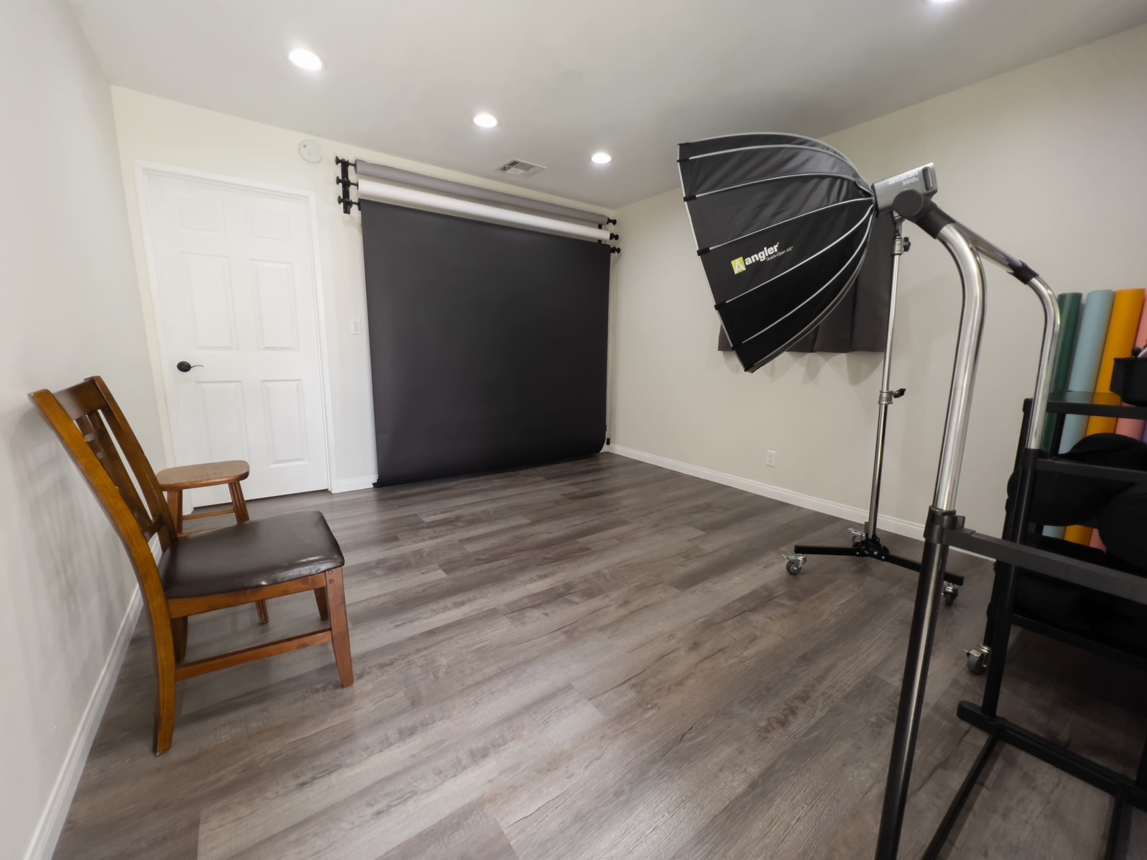 A photography studio features a wooden chair, a black backdrop, and a lighting umbrella on a hardwood floor.