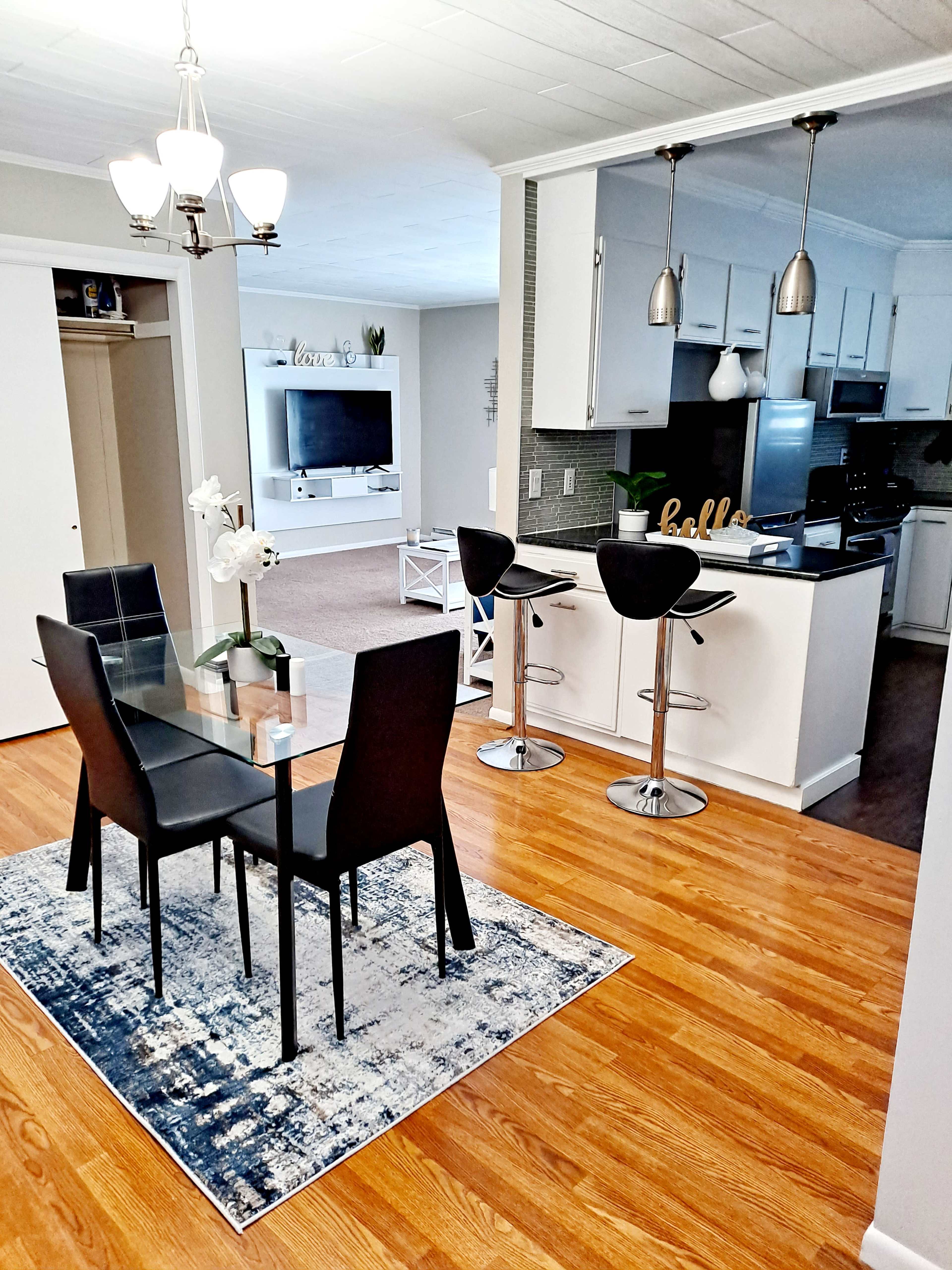 A modern kitchen and dining area features a glass dining table with black chairs, a rug, and bar stools at a breakfast counter.