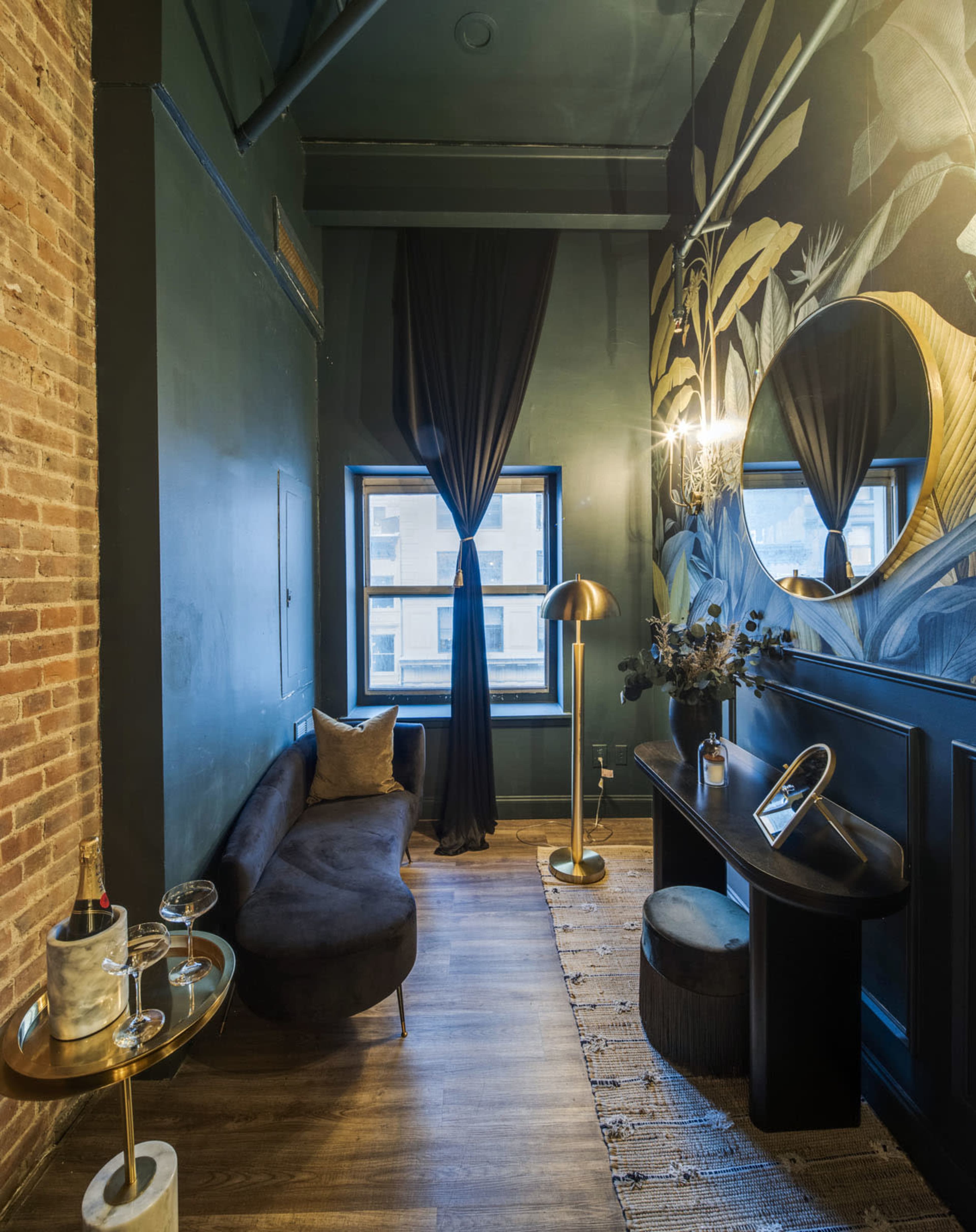 The image shows a narrow, stylish room with dark green walls, a large round mirror, a window, and a modern decor accentuated by plants and a cozy seating area.